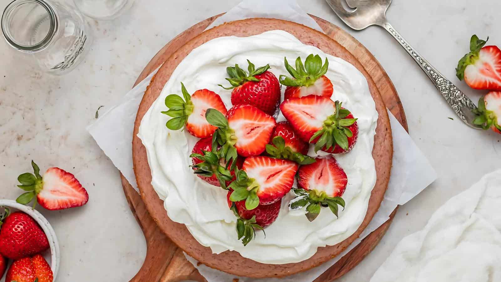 Round cake topped with a layer of whipped cream and fresh strawberries, sitting on a wooden board with a metal spoon and extra strawberries nearby.