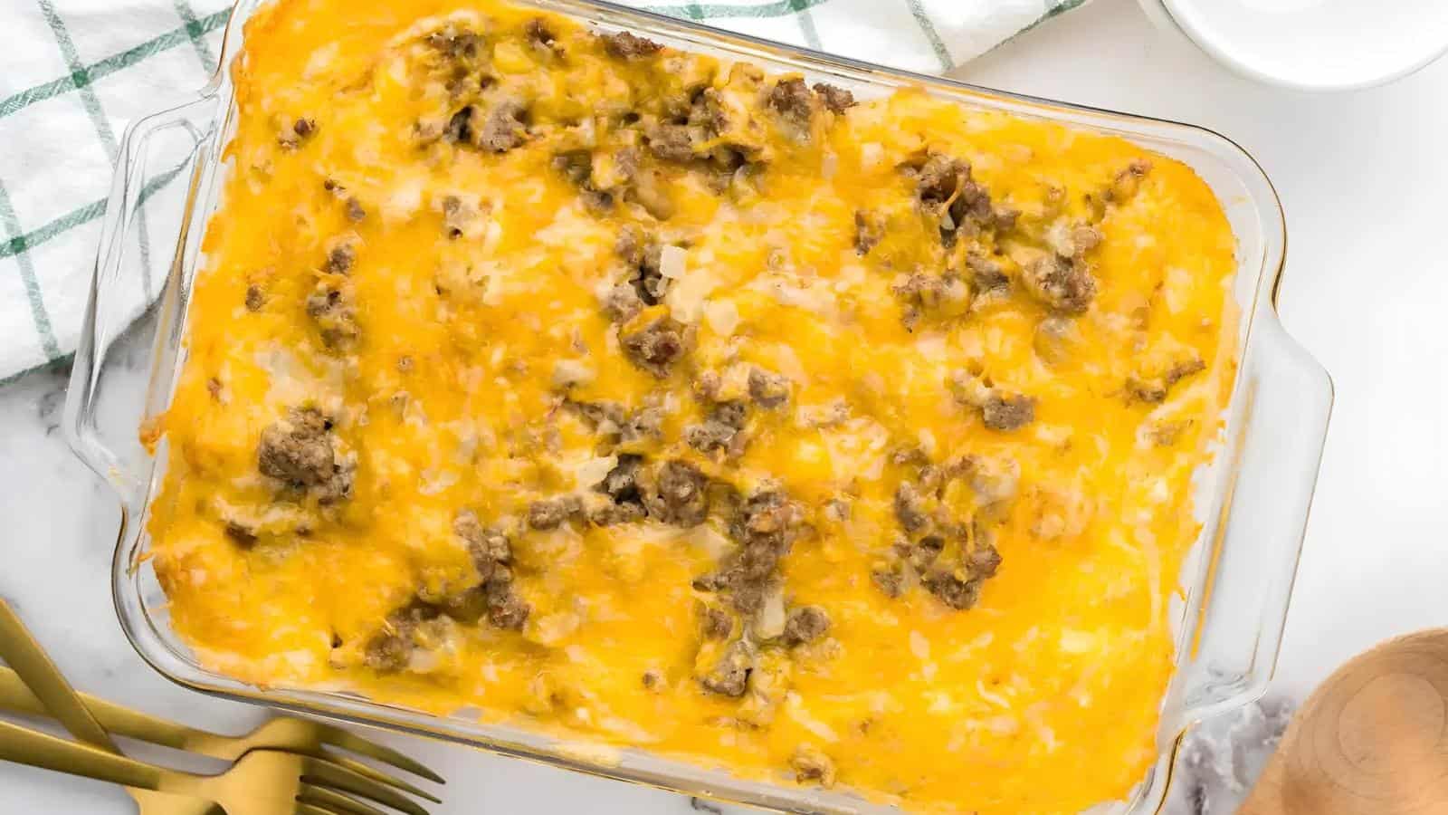 A casserole dish filled with a baked meal topped with melted cheese and crumbled sausage, set on a white surface with a striped cloth and gold forks nearby.