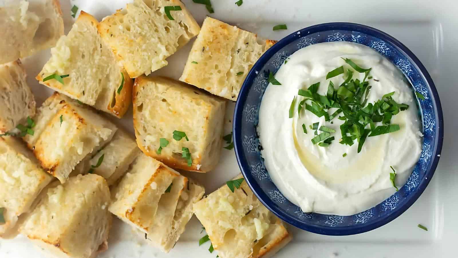 A plate with cubed pieces of bread next to a bowl of creamy white dip garnished with chopped herbs.