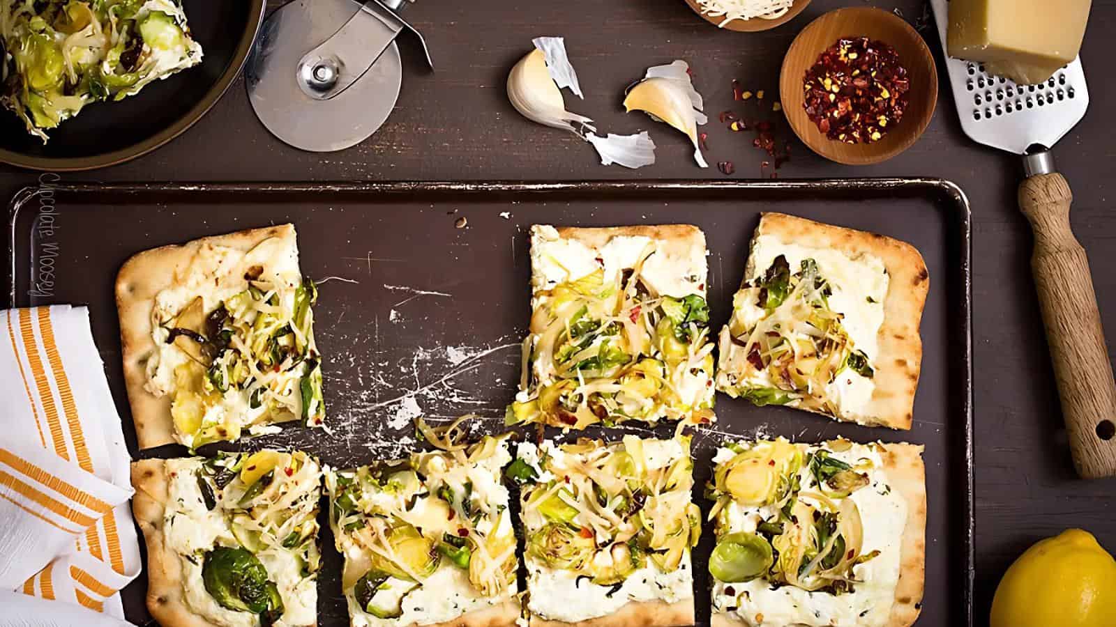 Rectangular flatbread pizza topped with cheese, Brussels sprouts, and onions is cut into six pieces on a baking sheet. Pizza cutter, garlic, red pepper flakes, and cheese are nearby.
