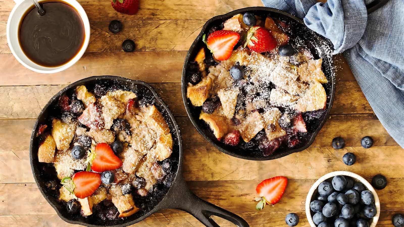Two cast iron pans with baked French toast topped with powdered sugar, strawberries, and blueberries on a wooden table. Nearby is a bowl of blueberries, a mug of coffee, and a blue cloth.
