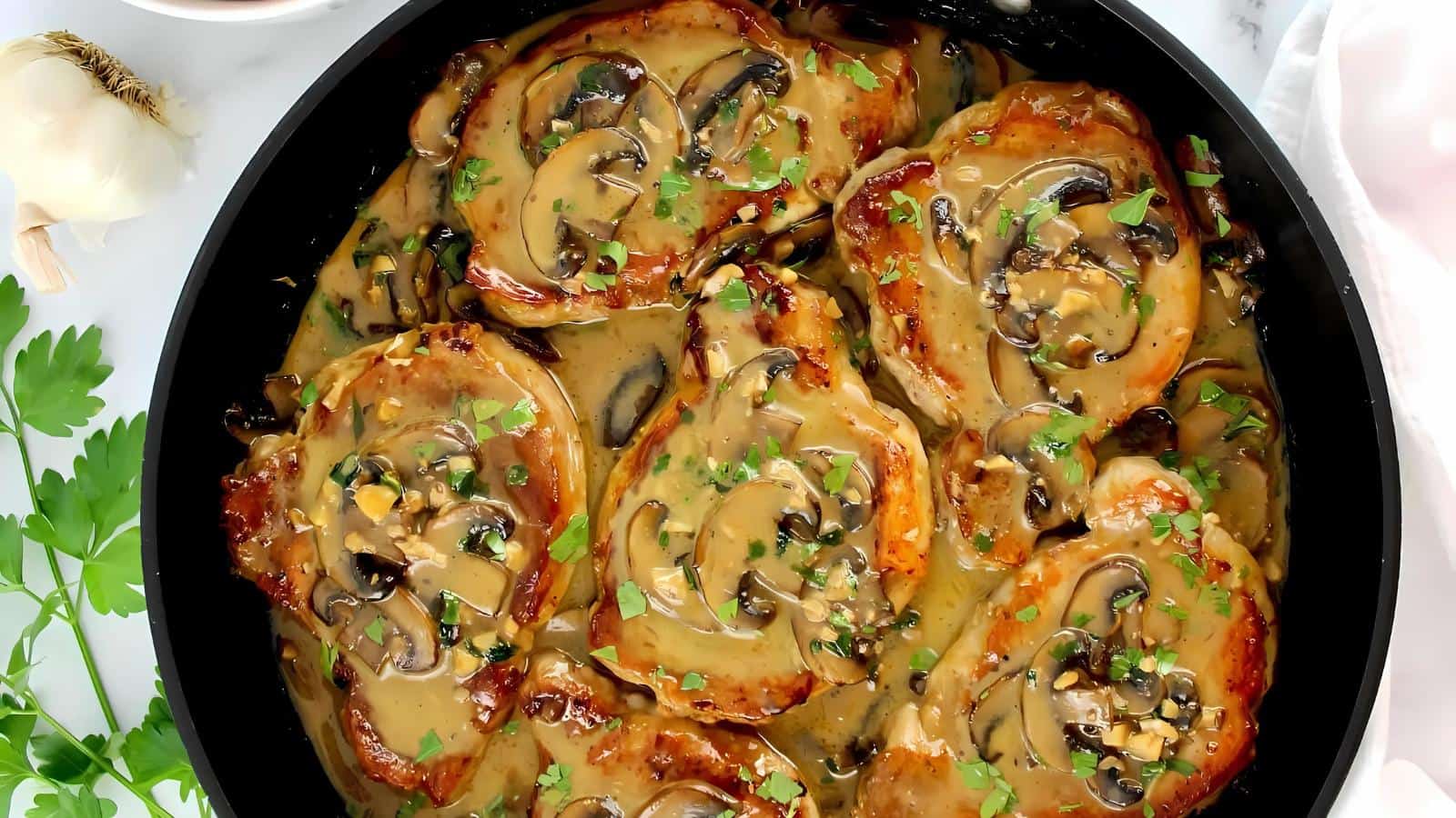 Pork chops cooked in a creamy mushroom sauce, garnished with chopped herbs, served in a black skillet.