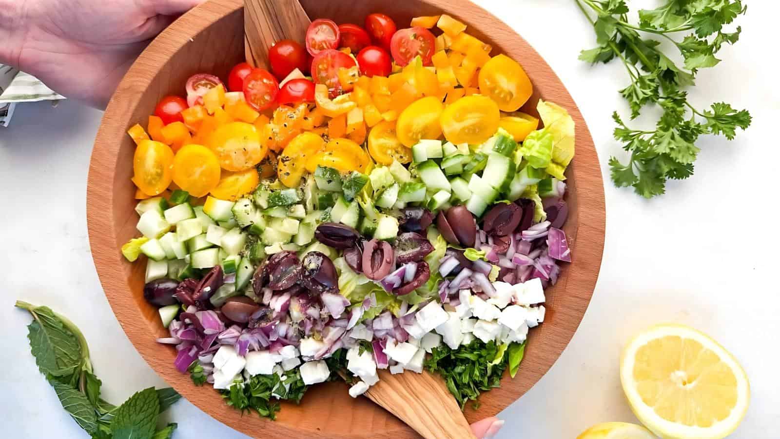 A wooden bowl filled with a colorful chopped salad, including cherry tomatoes, yellow peppers, cucumbers, olives, red onions, feta cheese, and herbs.