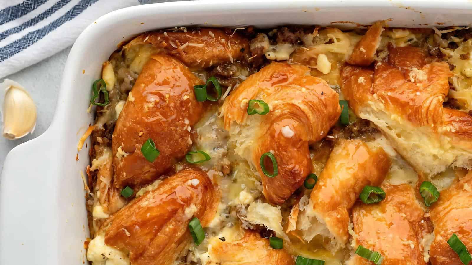 Baked casserole dish with croissants, melted cheese, and green onion slices on top. A striped cloth and garlic clove are visible in the background.