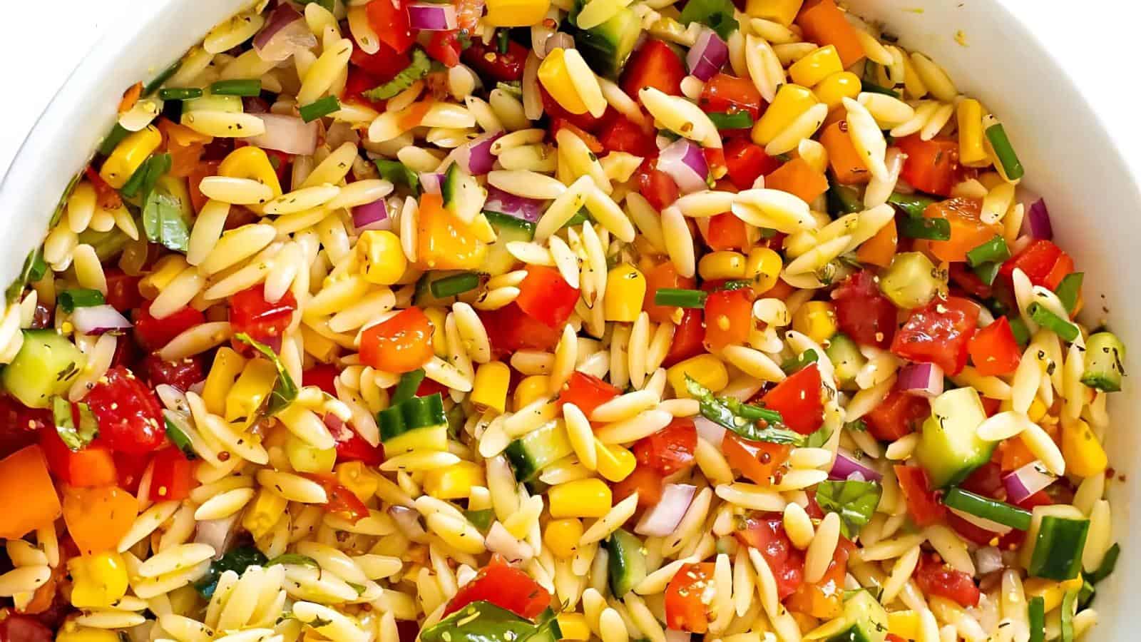 A close-up of a bowl filled with orzo pasta salad mixed with chopped cucumbers, tomatoes, red onions, bell peppers, corn, and herbs.
