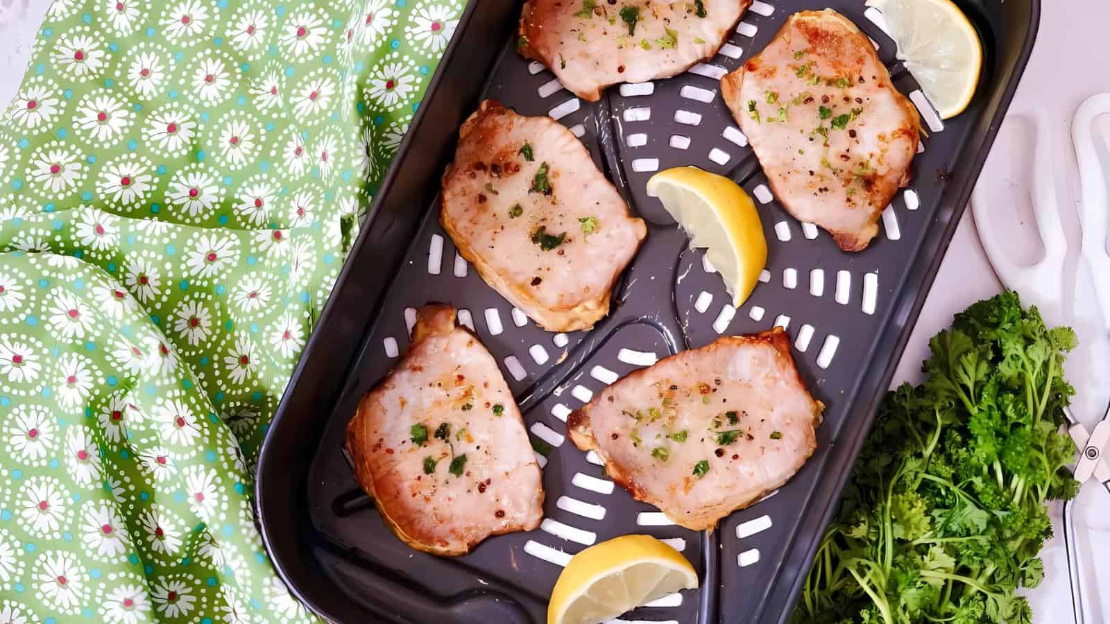 Five seasoned pork chops on a baking tray with lemon wedges, surrounded by a green patterned cloth and fresh parsley.