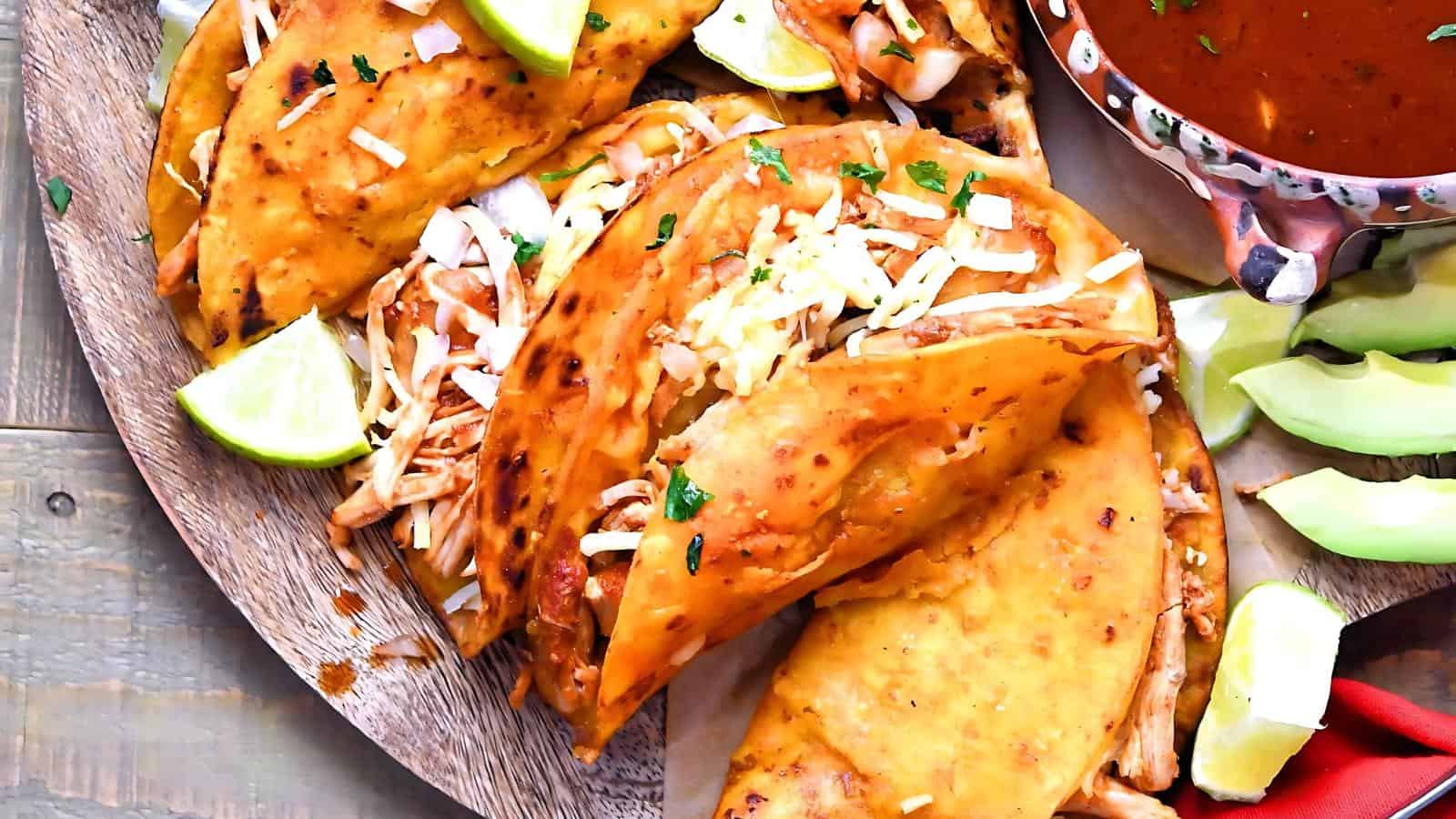 Three crispy tacos filled with shredded chicken and cheese are served on a wooden plate with lime wedges on the side and a bowl of sauce partially visible.