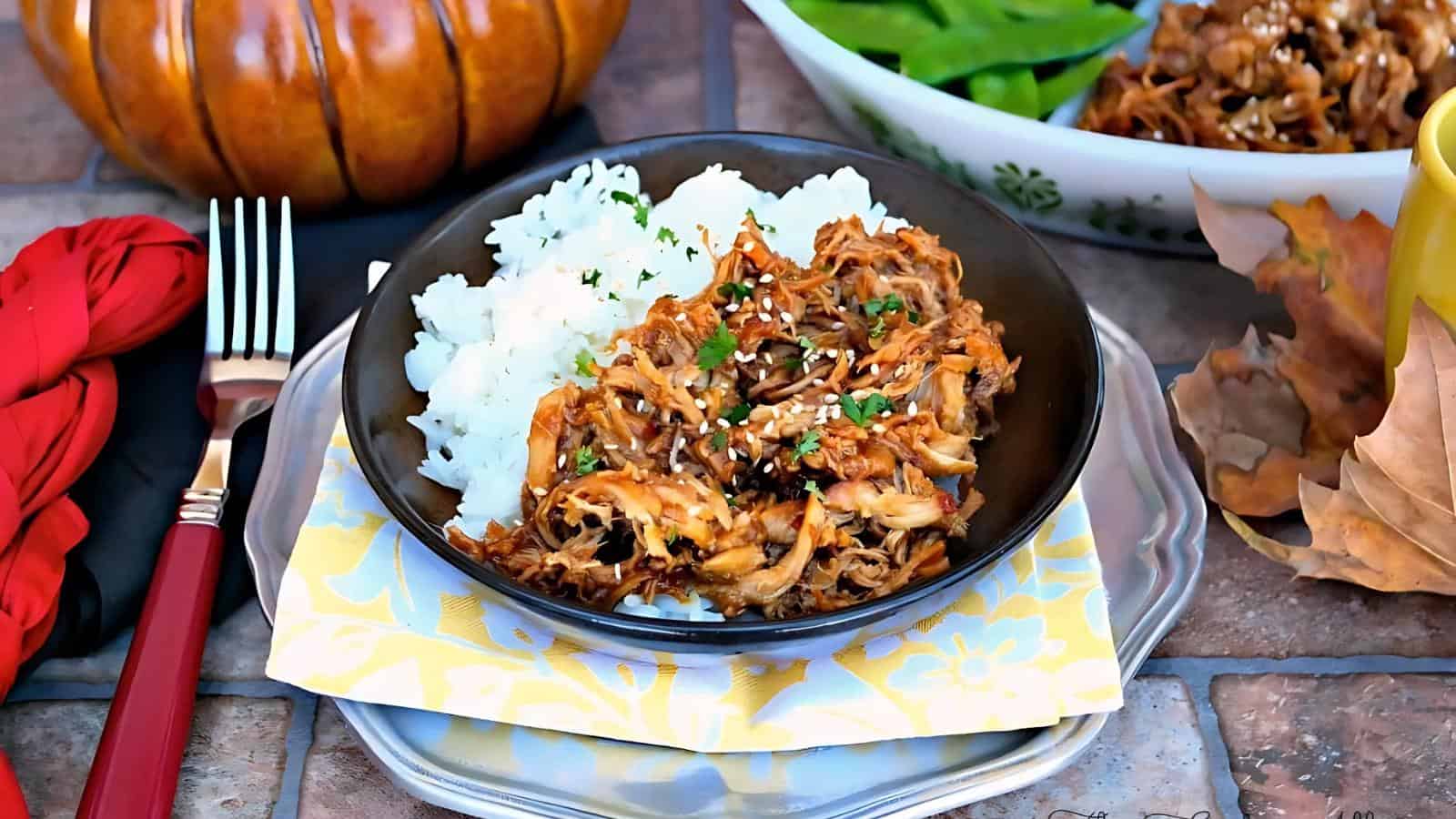 A black bowl filled with shredded meat topped with sesame seeds and chopped herbs, served with white rice, placed on a stack of plates with a napkin, beside autumn decor.