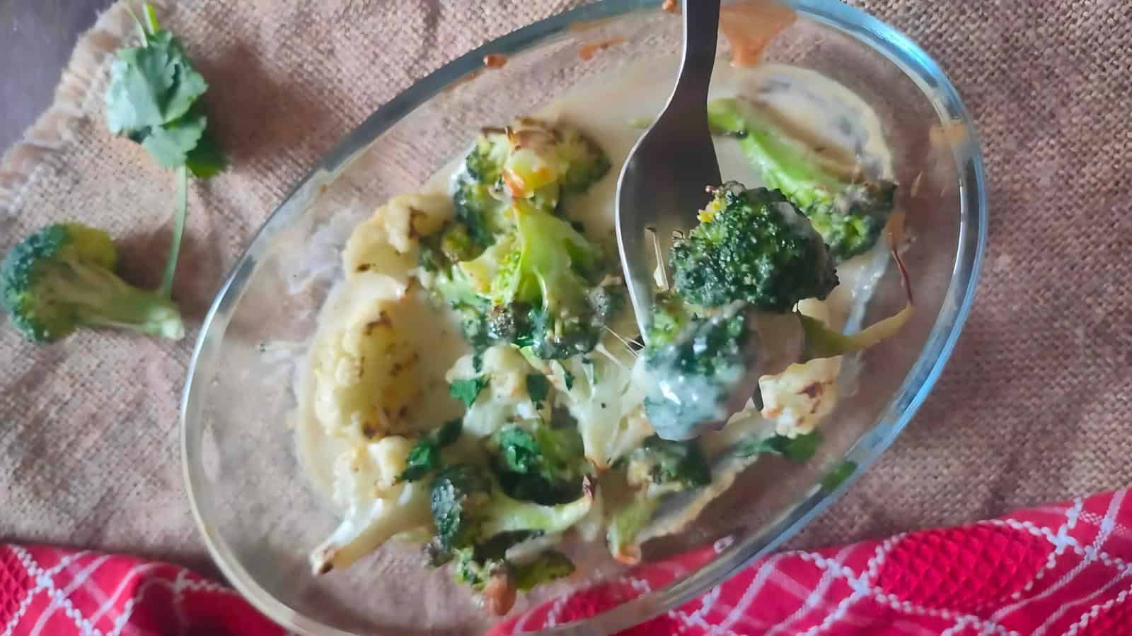 A glass dish with baked broccoli and cauliflower in a creamy sauce; a fork holds a portion above the dish. A parsley sprig and red towel are nearby.