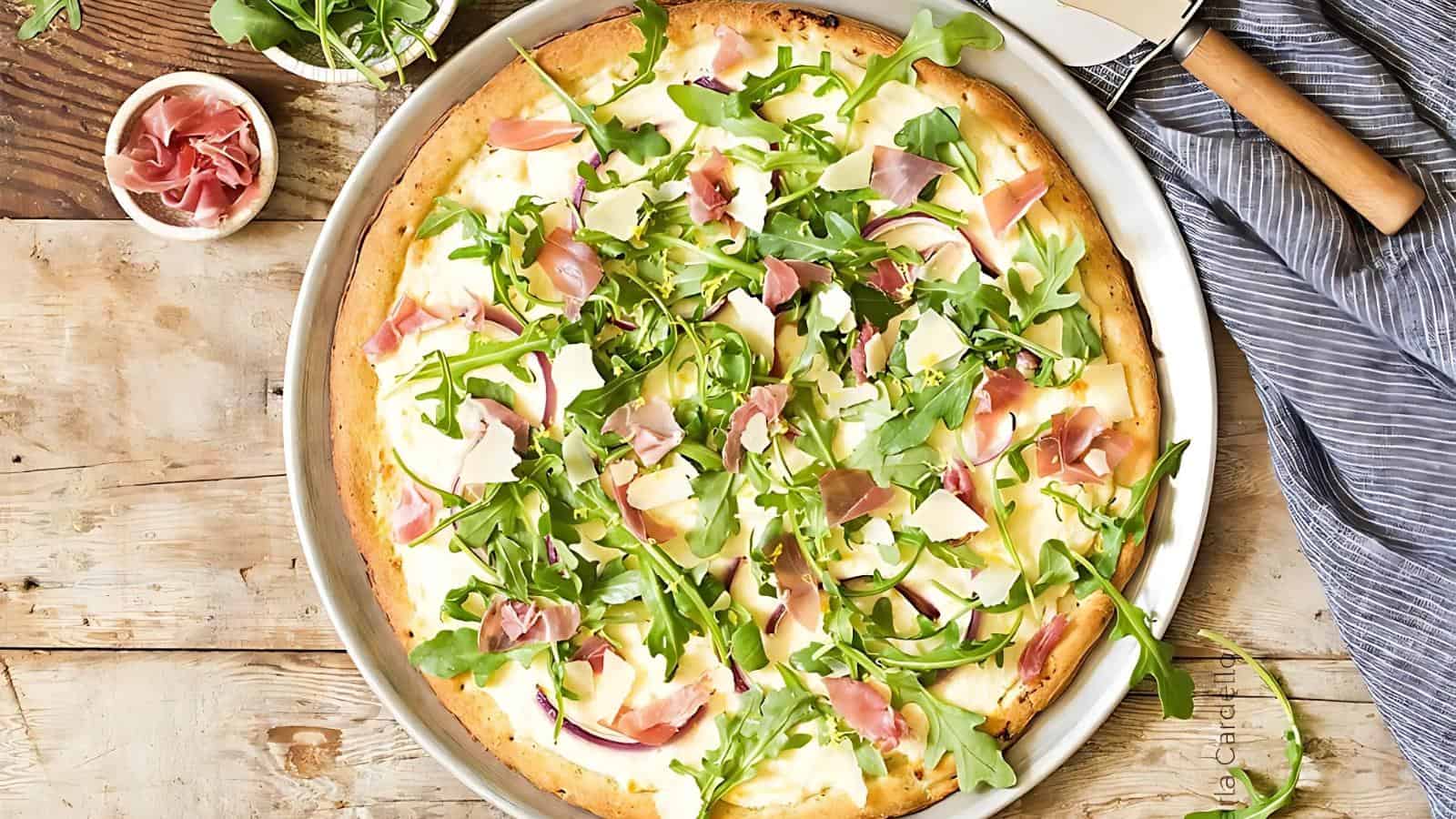 Thin crust pizza topped with arugula, prosciutto, shaved cheese, and red onion slices on a round metal tray, with a pizza cutter and napkin nearby.