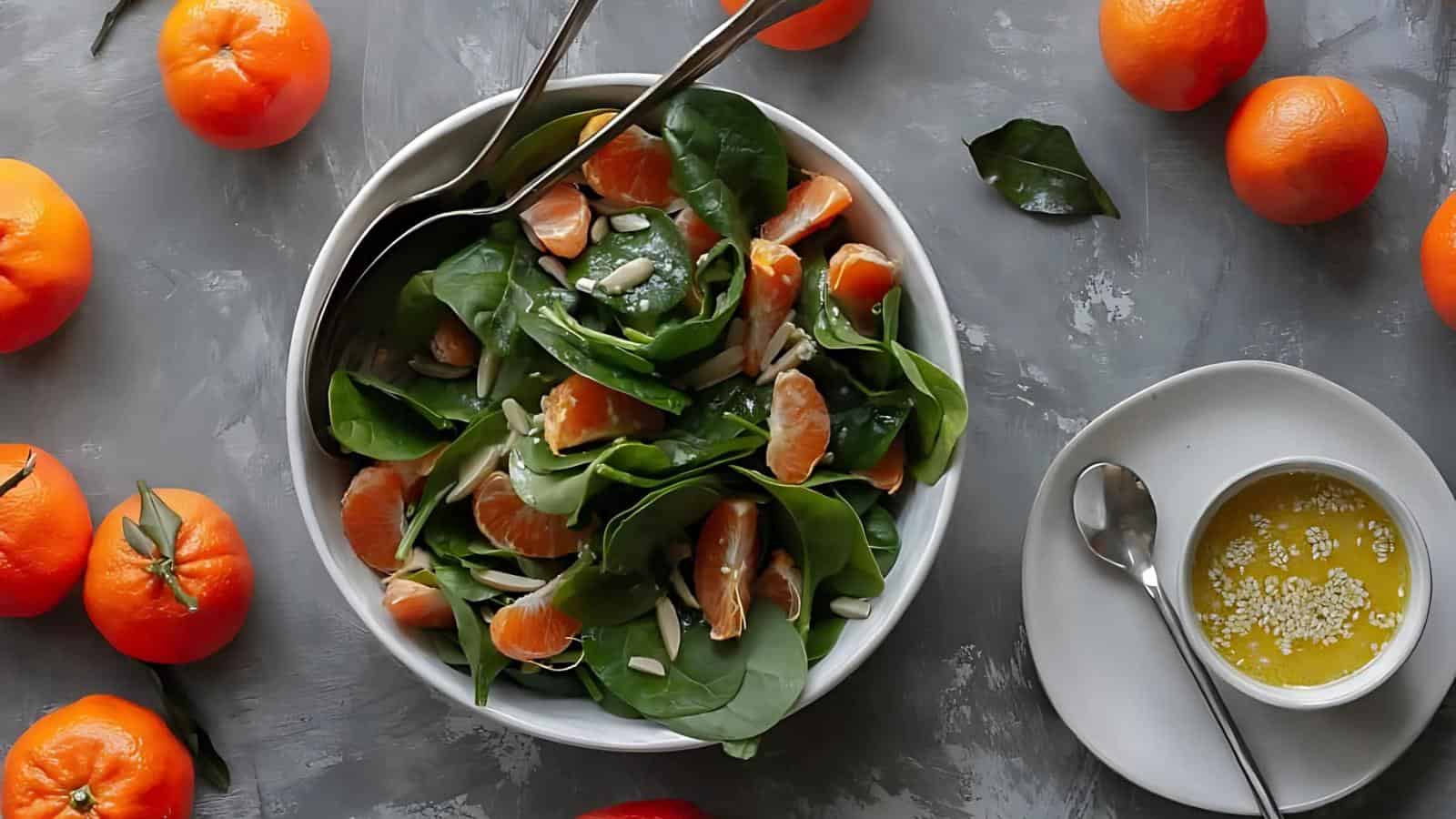 A bowl of spinach and mandarin orange salad with serving utensils, surrounded by whole mandarins, next to a plate with a cup of dressing on a gray surface.