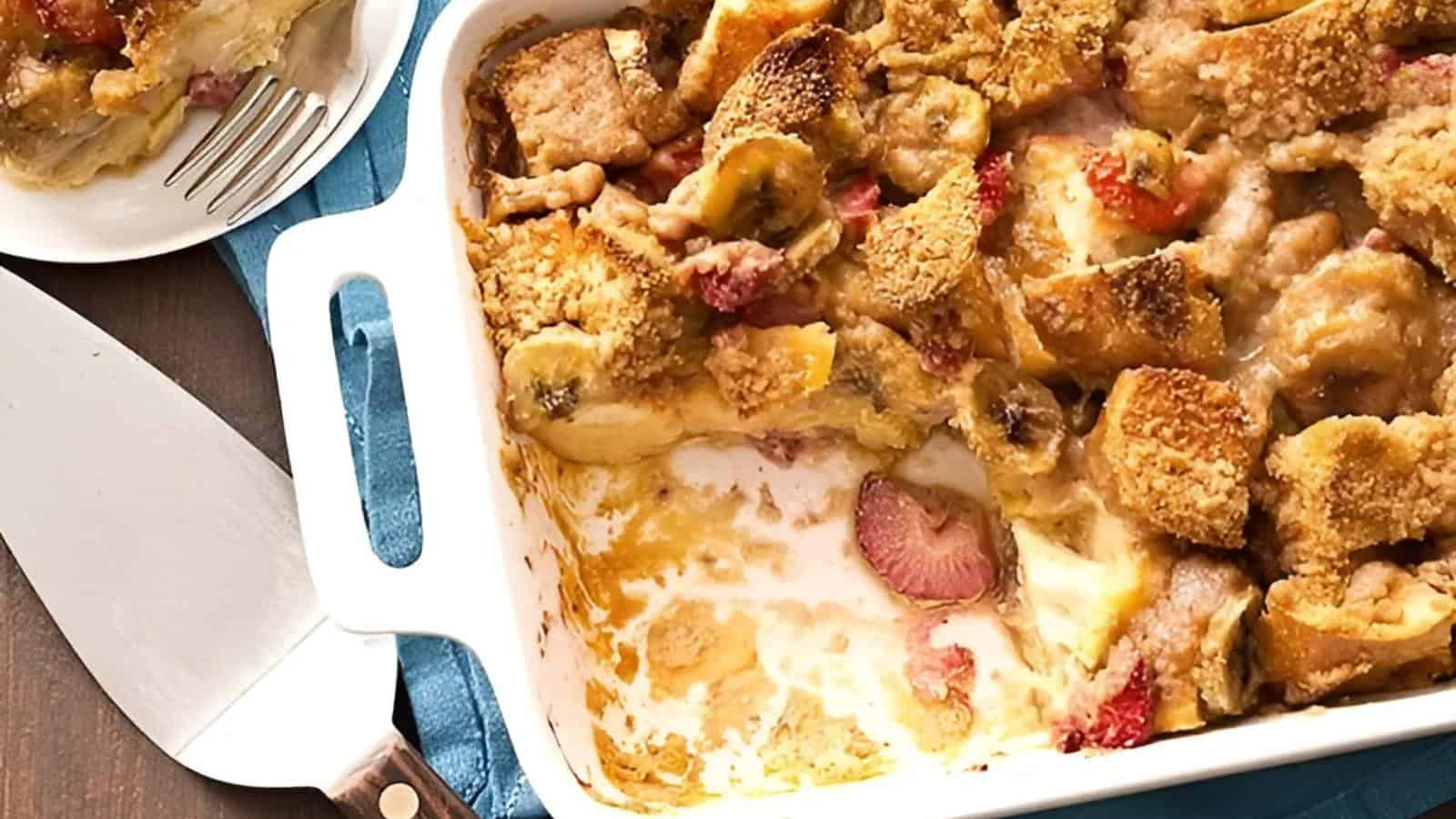 Baked dish of bread, strawberries, and bananas in a creamy sauce, partially served onto a plate beside a serving utensil.