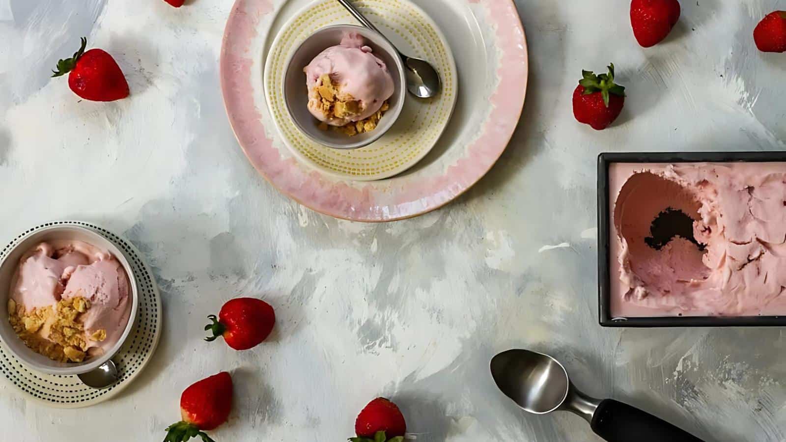 Two bowls of strawberry ice cream with crumbled topping, a rectangular container of ice cream, ice cream scoop, and scattered strawberries on a textured surface.