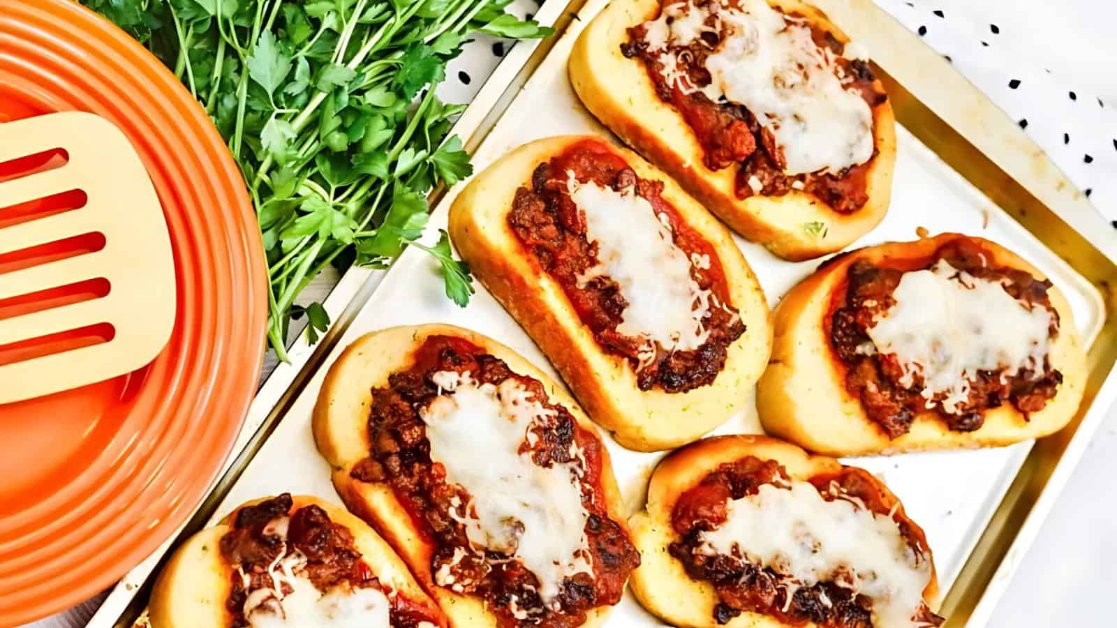 Six mini pizzas with melted cheese and tomato-based topping are arranged on a baking tray next to a bunch of fresh parsley and orange plates with a matching spatula.