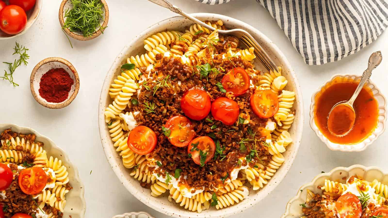 A bowl of rotini pasta topped with a tomato-based sauce, ground meat, cherry tomato halves, and fresh herbs, surrounded by small bowls of seasonings and sauce on a white table.
