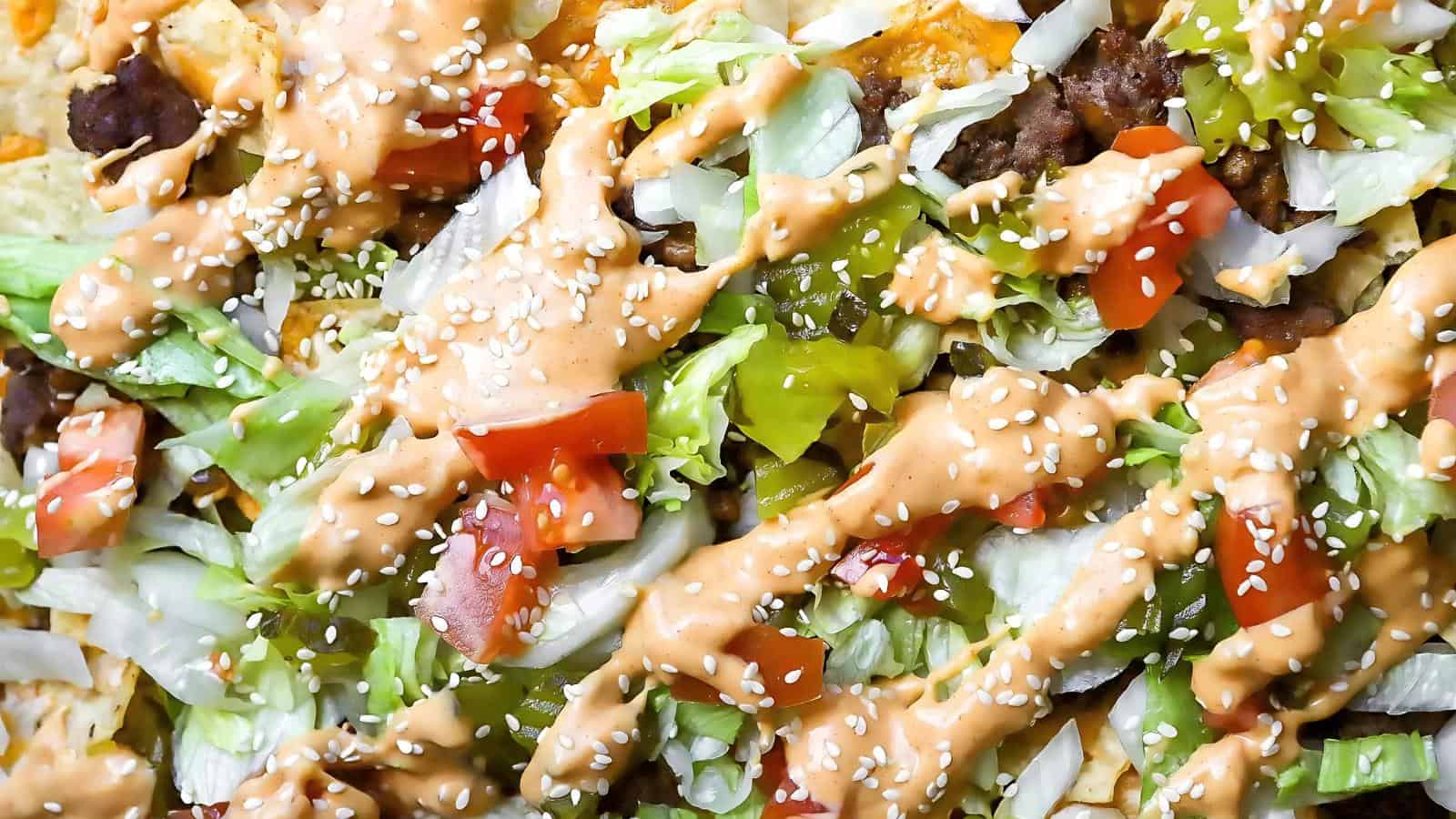 Close-up of nachos topped with ground meat, chopped lettuce, diced tomatoes, pickles, sesame seeds, and a drizzle of creamy sauce.