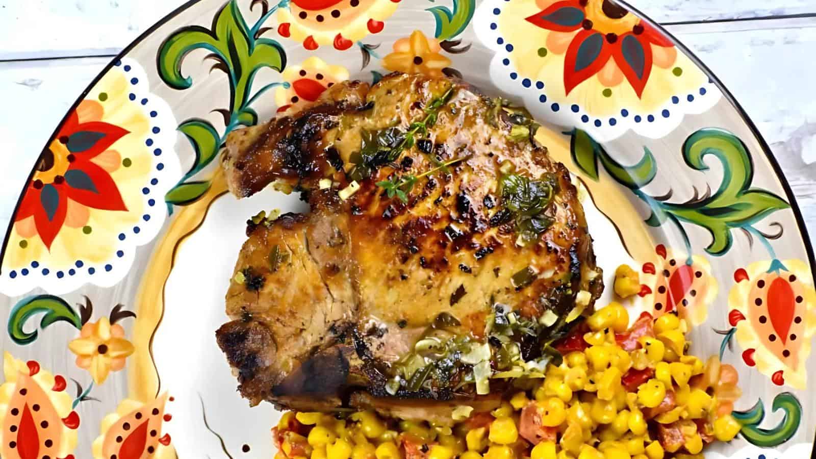 Grilled pork chop with herbs served on a decorative plate, accompanied by a side of corn and diced vegetables.