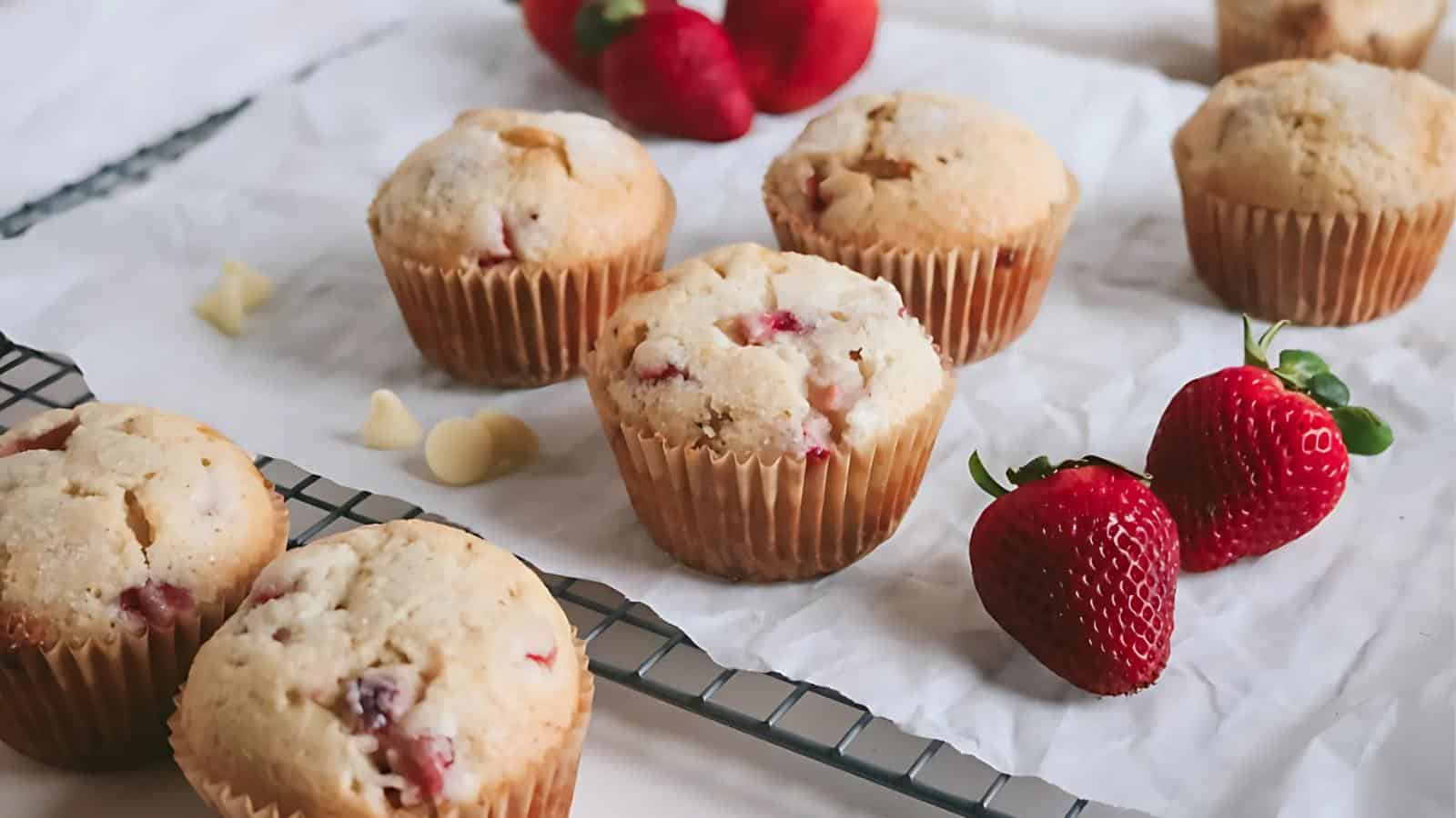 Strawberry muffins on a cooling rack with fresh strawberries and white chocolate chips on a parchment-lined surface.