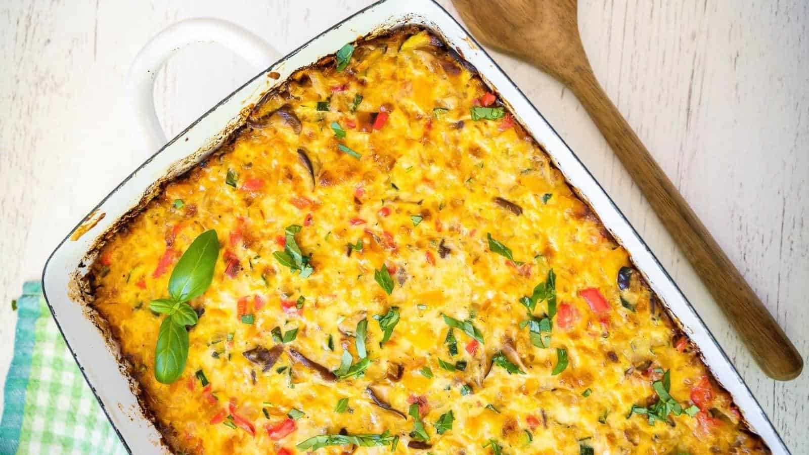 Baked casserole with melted cheese and vegetables in a white dish, accompanied by a wooden spoon on a light surface.