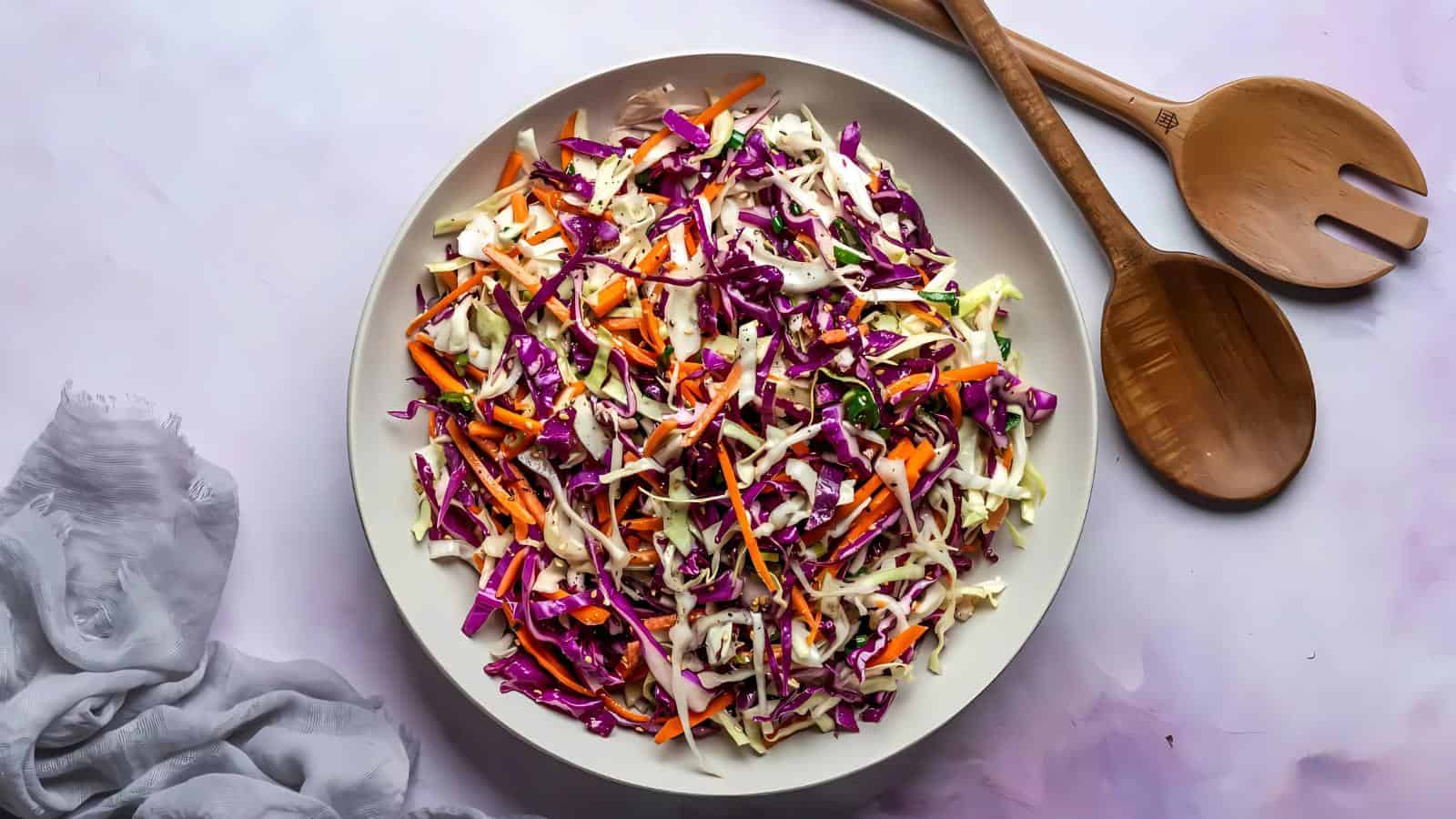 A bowl of coleslaw made with shredded red and green cabbage and carrots, placed next to wooden salad servers on a light surface.