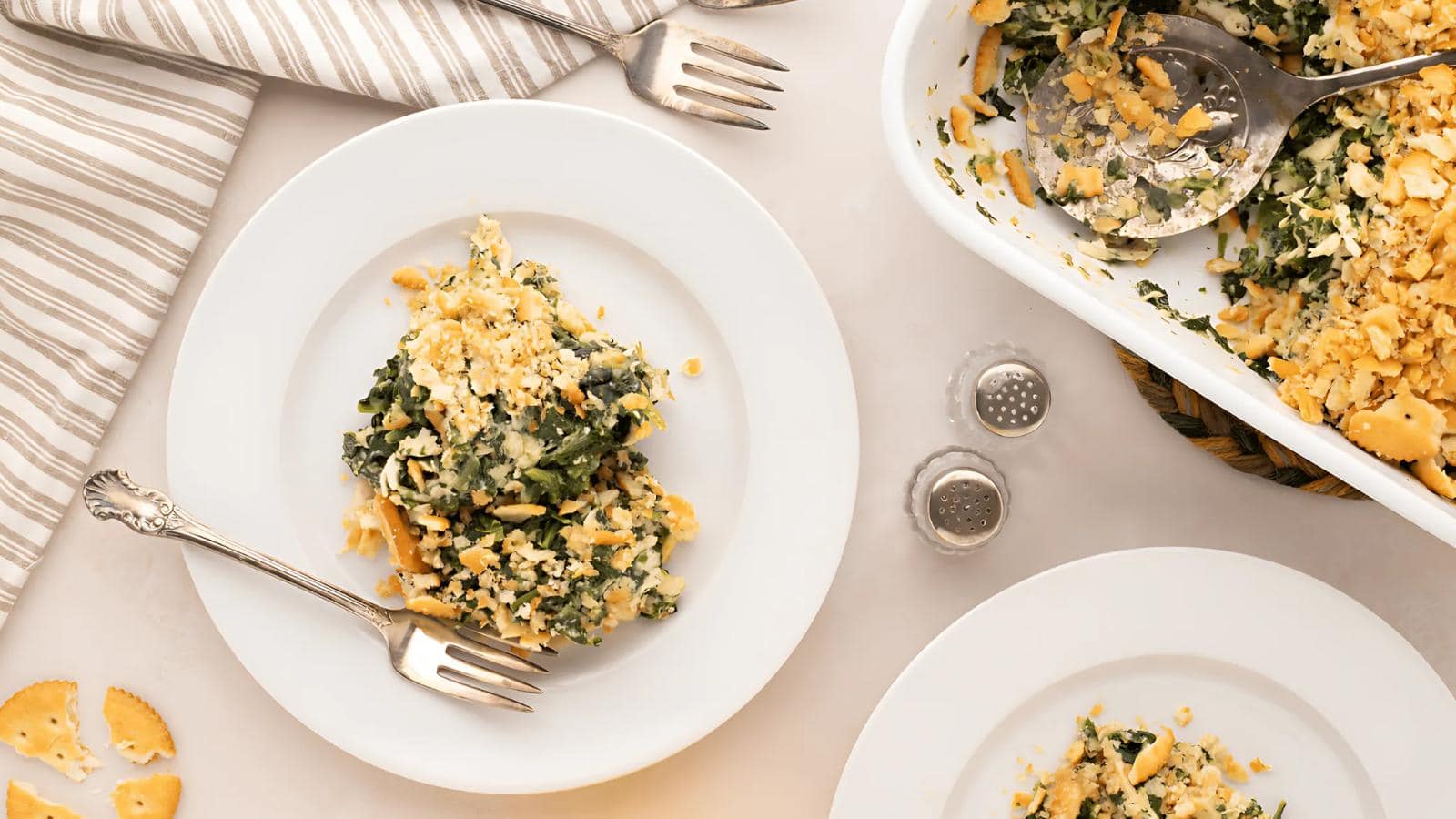 A serving of spinach casserole with a crumb topping on a white plate, shown with utensils and a partially empty casserole dish nearby.