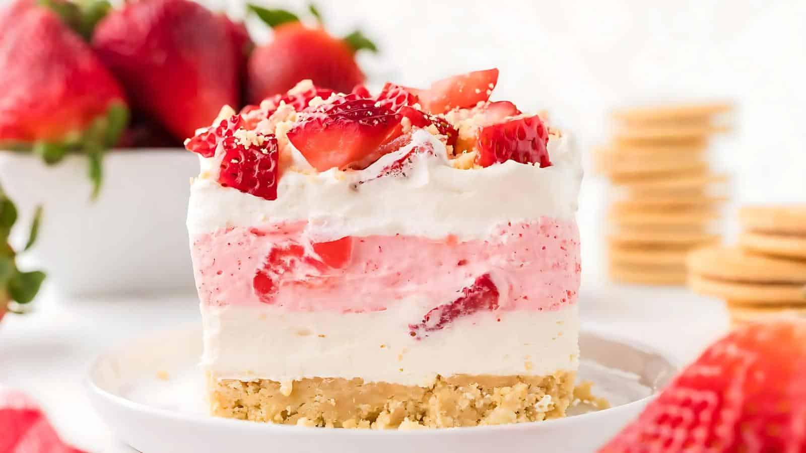 A slice of layered strawberry dessert with a crumbly crust, creamy layers, and fresh strawberries on top, served on a white plate.