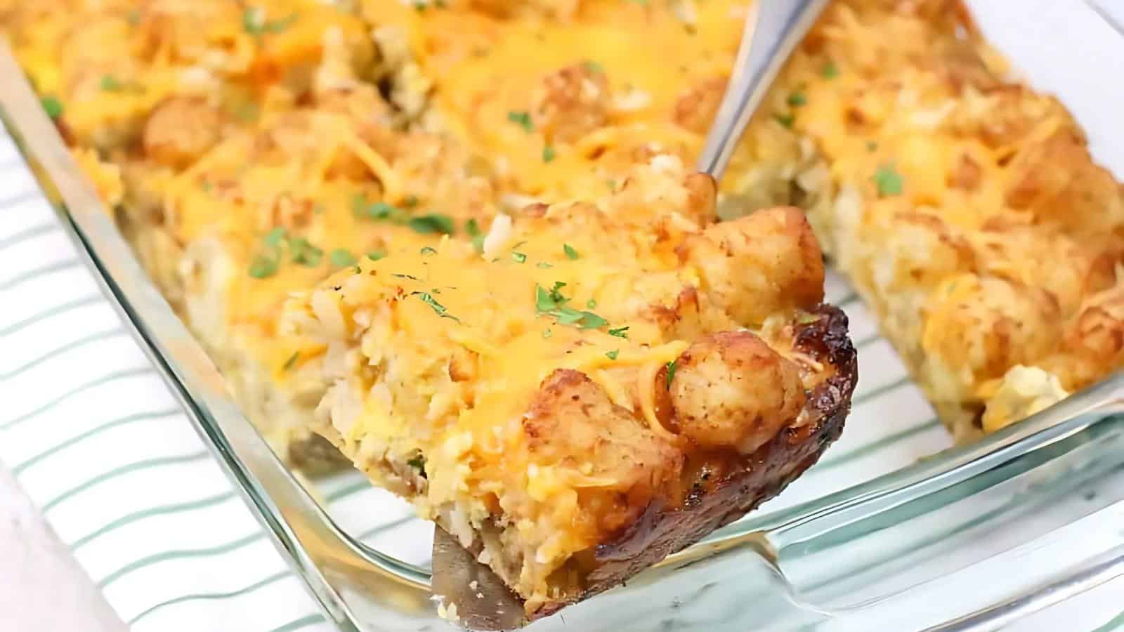 A serving of tater tot casserole being lifted from a glass baking dish, showing layers of cheese and garnished with parsley.