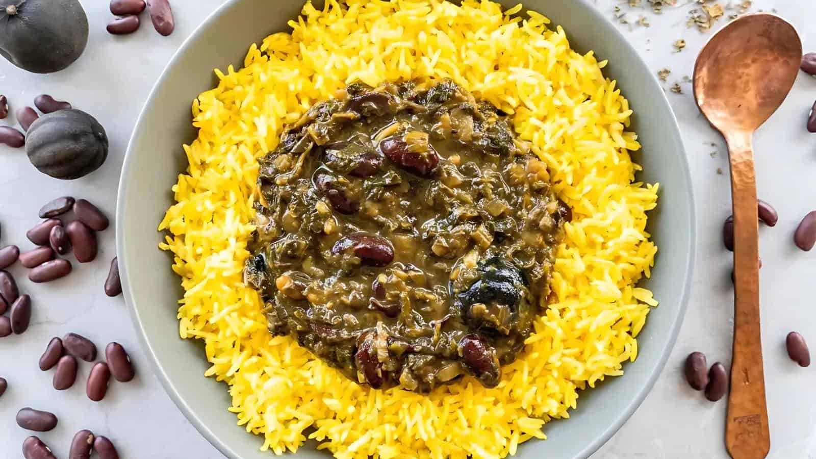 A bowl of yellow rice topped with a green vegetable and red kidney bean stew, surrounded by scattered beans and dried limes, with a copper spoon on the side.
