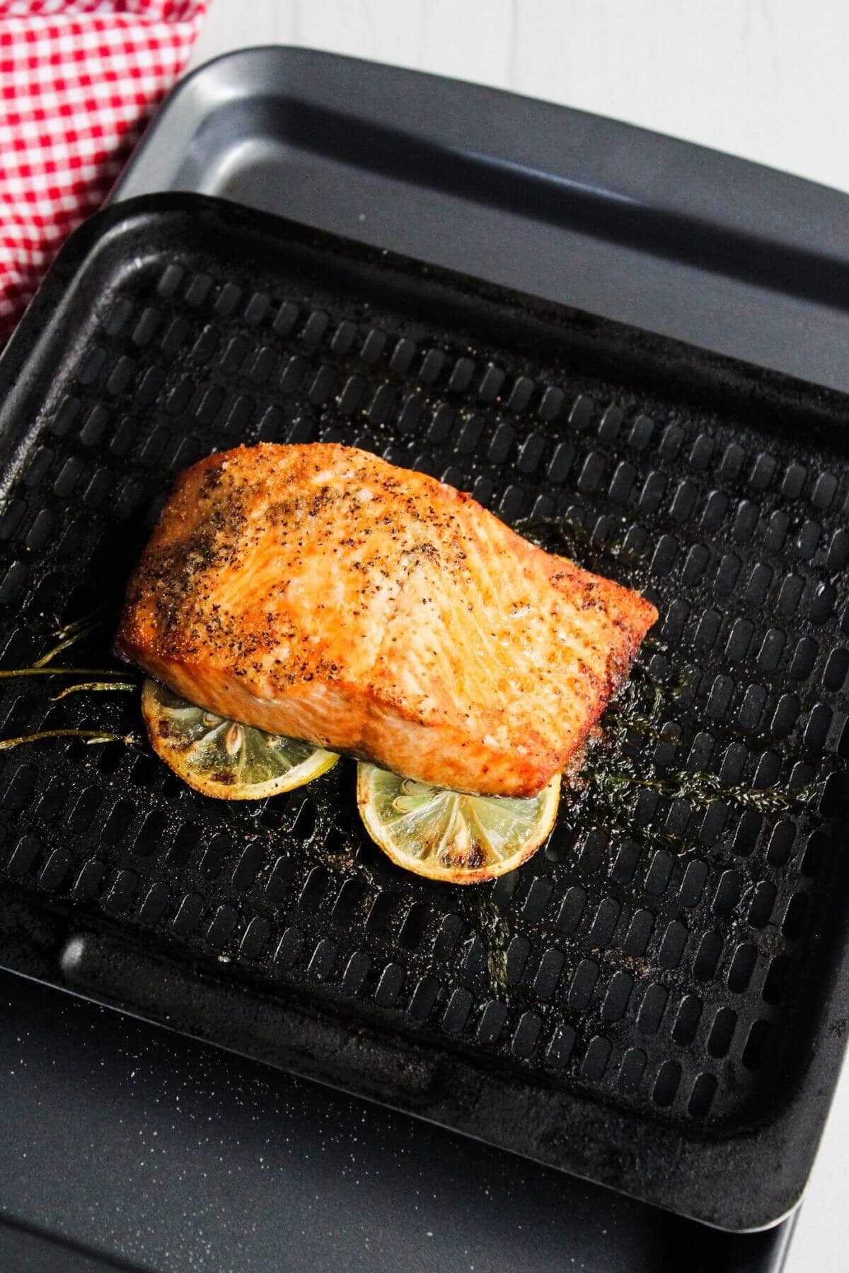 Grilled salmon fillet with pepper seasoning on a black grill pan, resting on lemon slices, with a red and white checkered cloth nearby.