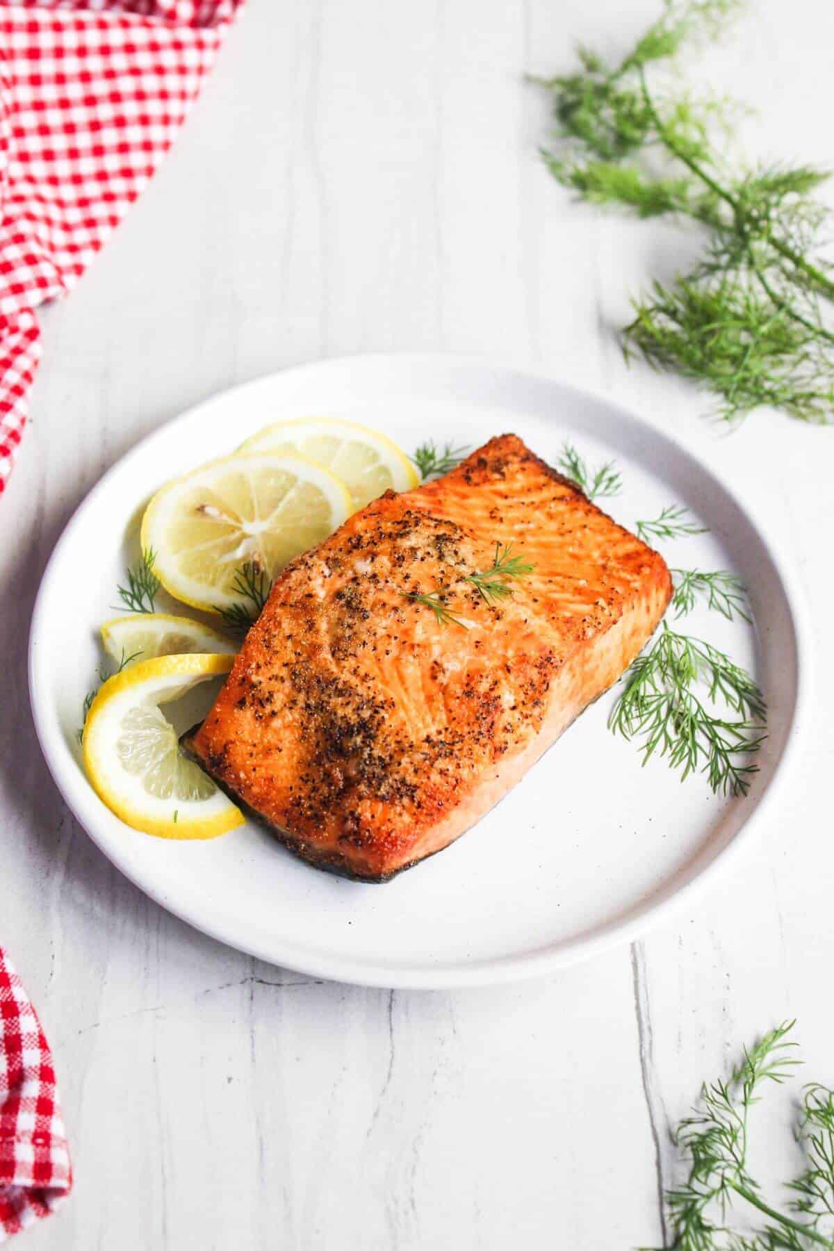 Grilled salmon fillet on a plate with lemon slices and dill garnish, placed on a white wooden surface with a red checkered napkin nearby.
