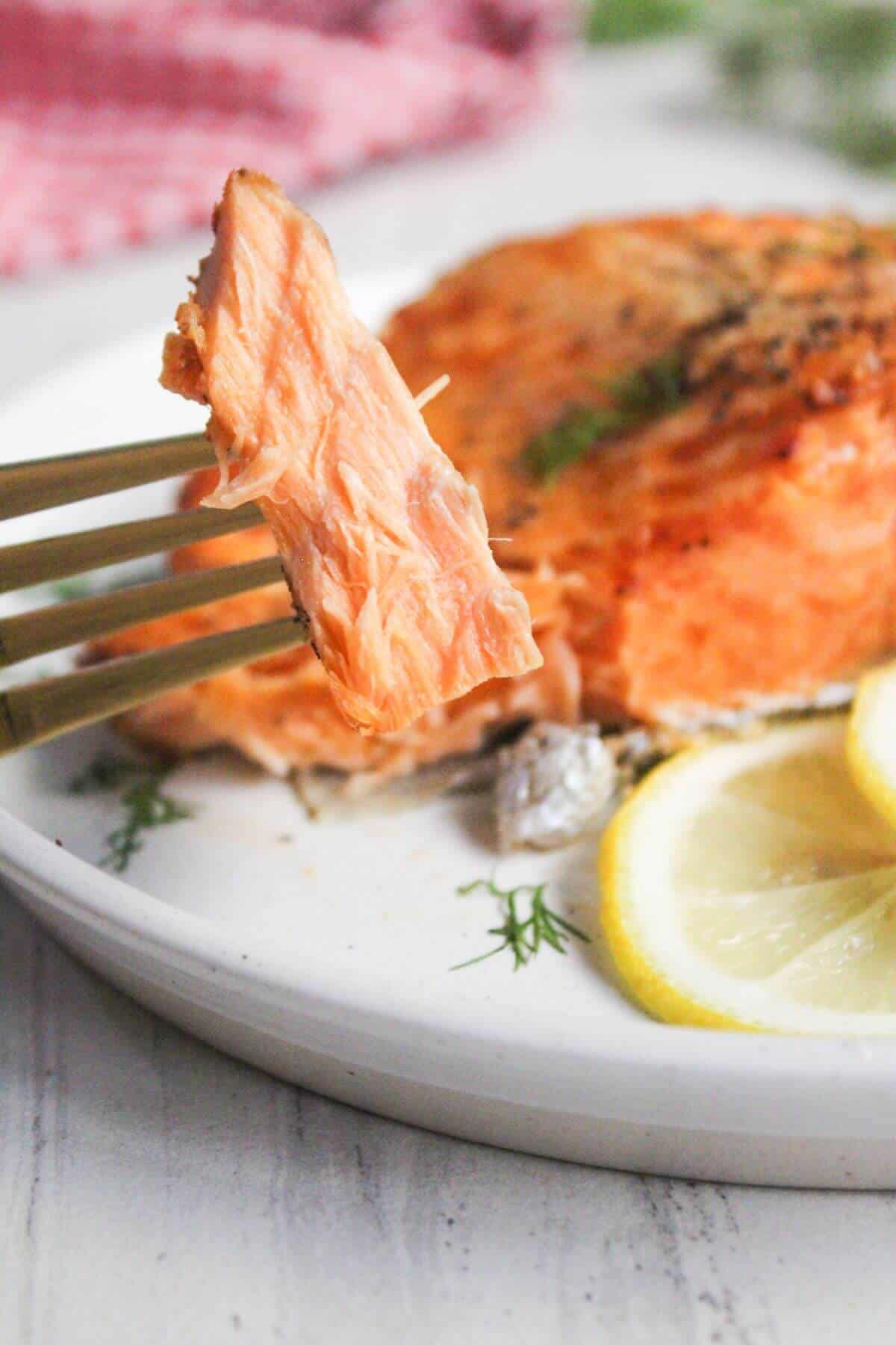 A fork holding a piece of cooked salmon over a plate with salmon, garnished with dill and lemon slices.