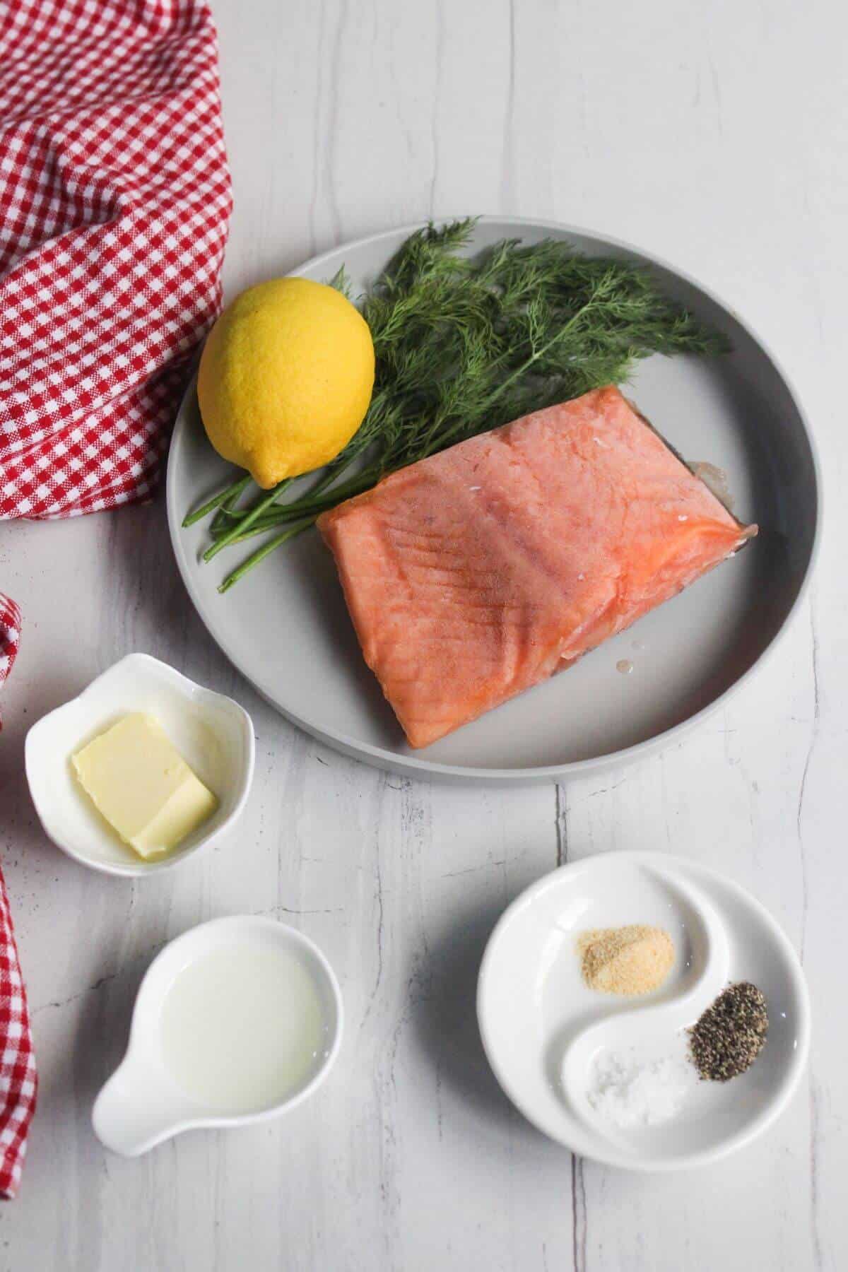 Salmon fillet on a plate with dill and a lemon. Nearby are small dishes with butter, milk, and seasoning. A red checkered cloth is partially visible.