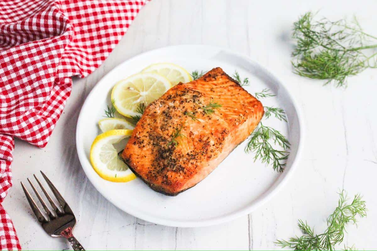 Grilled salmon fillet on a white plate with lemon slices and dill sprigs. A red checkered cloth and a fork are nearby on a white surface.