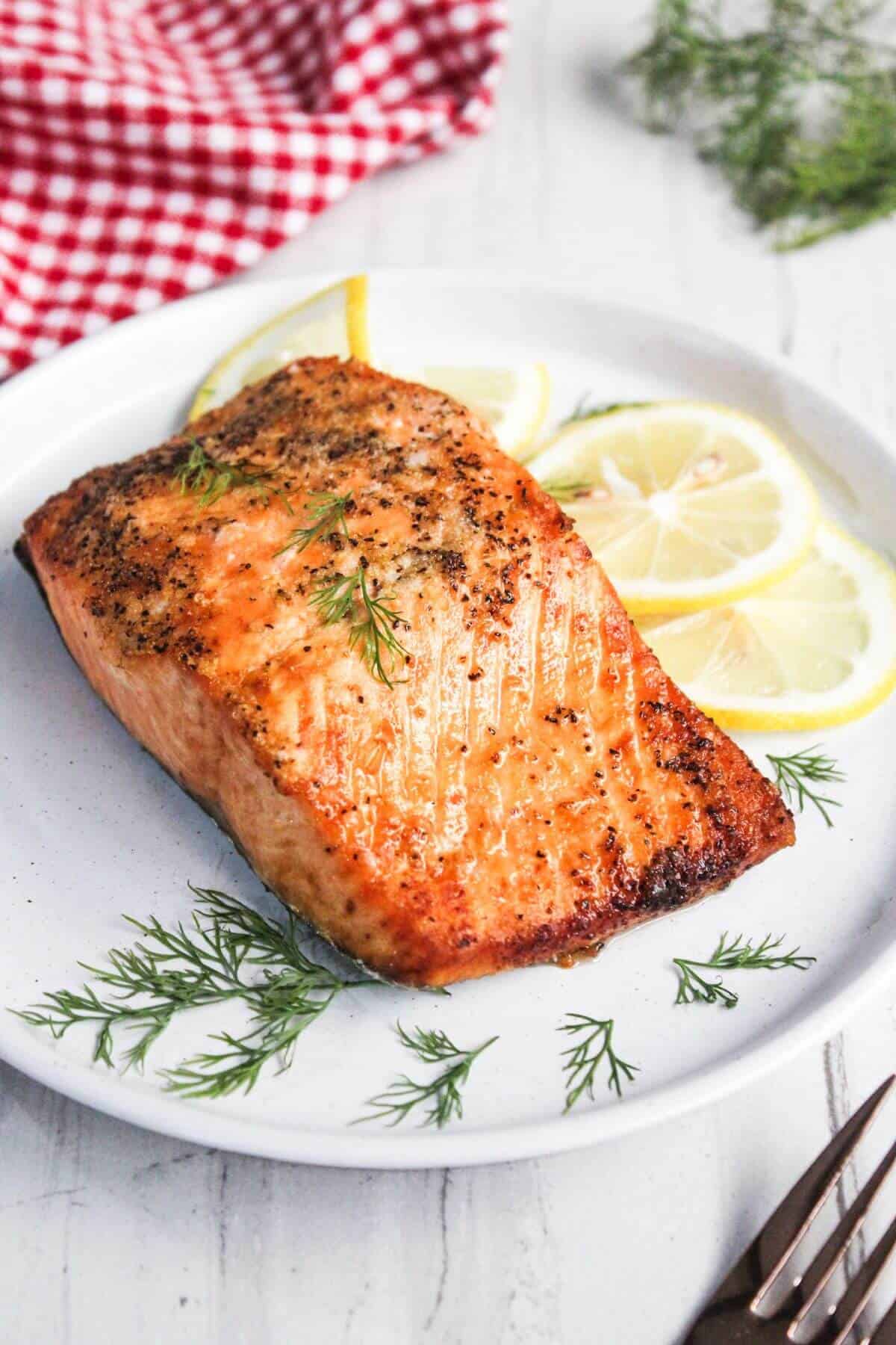 Grilled salmon fillet garnished with fresh dill and lemon slices on a white plate, with a red checkered cloth partially visible in the background.