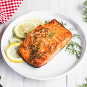 Grilled salmon fillet on a white plate, garnished with lemon slices and dill. A red-checkered cloth is partially visible beside the plate.