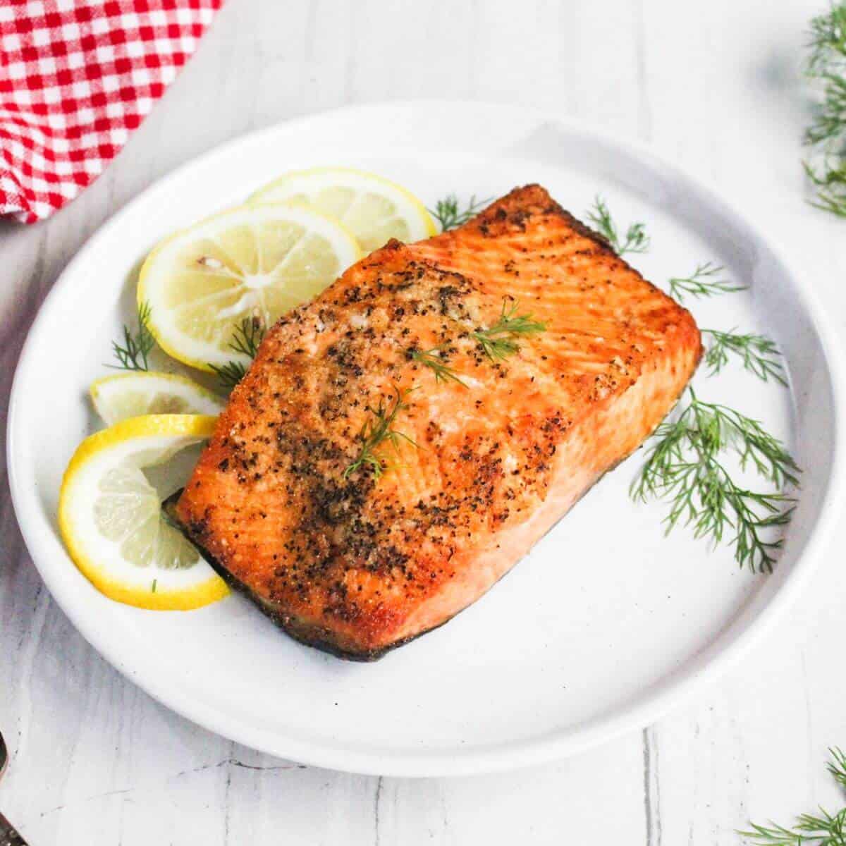 Grilled salmon fillet on a white plate, garnished with lemon slices and dill. A red-checkered cloth is partially visible beside the plate.