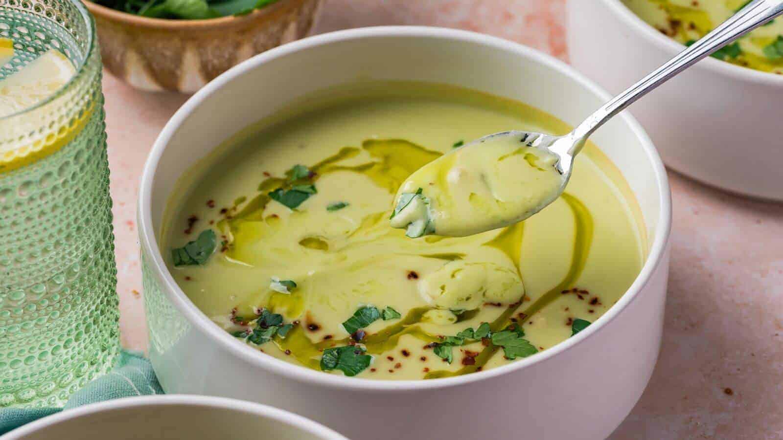 A bowl of creamy green soup garnished with herbs and oil, with a spoon lifting some soup from the bowl.