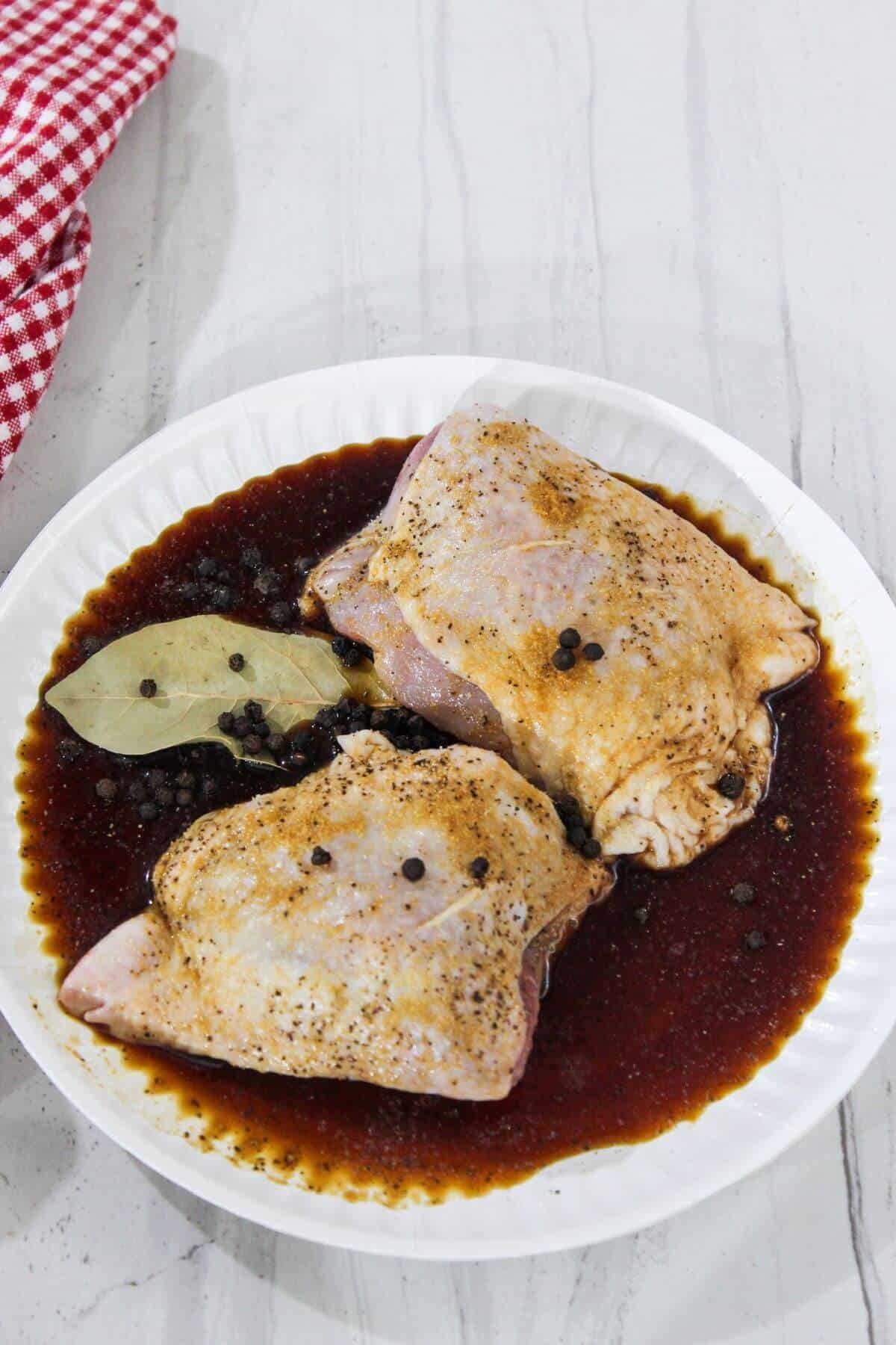Two raw chicken thighs seasoned with peppercorns and a bay leaf on a white plate, surrounded by a dark marinade. A red and white checkered cloth is partially visible on the left.