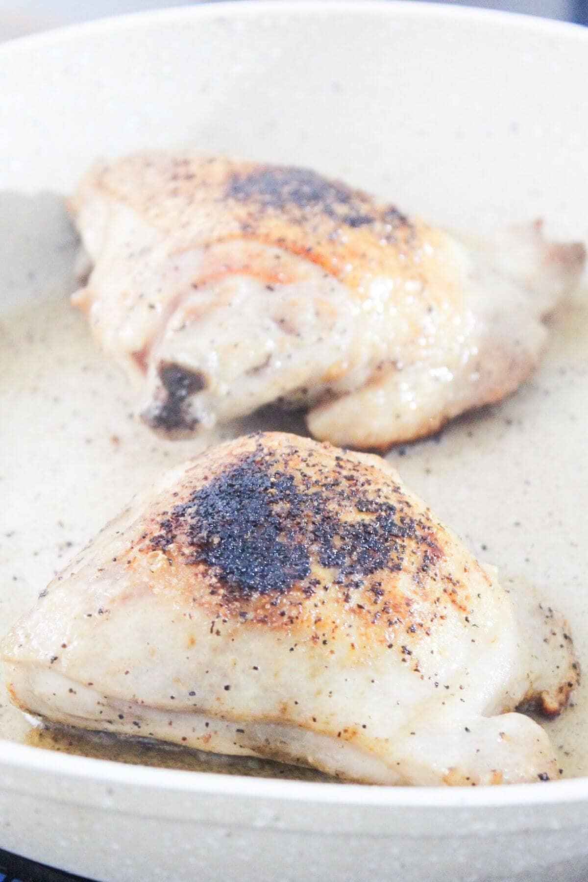 Two seasoned chicken thighs are cooking in a pan, showing a slightly browned and crispy skin.