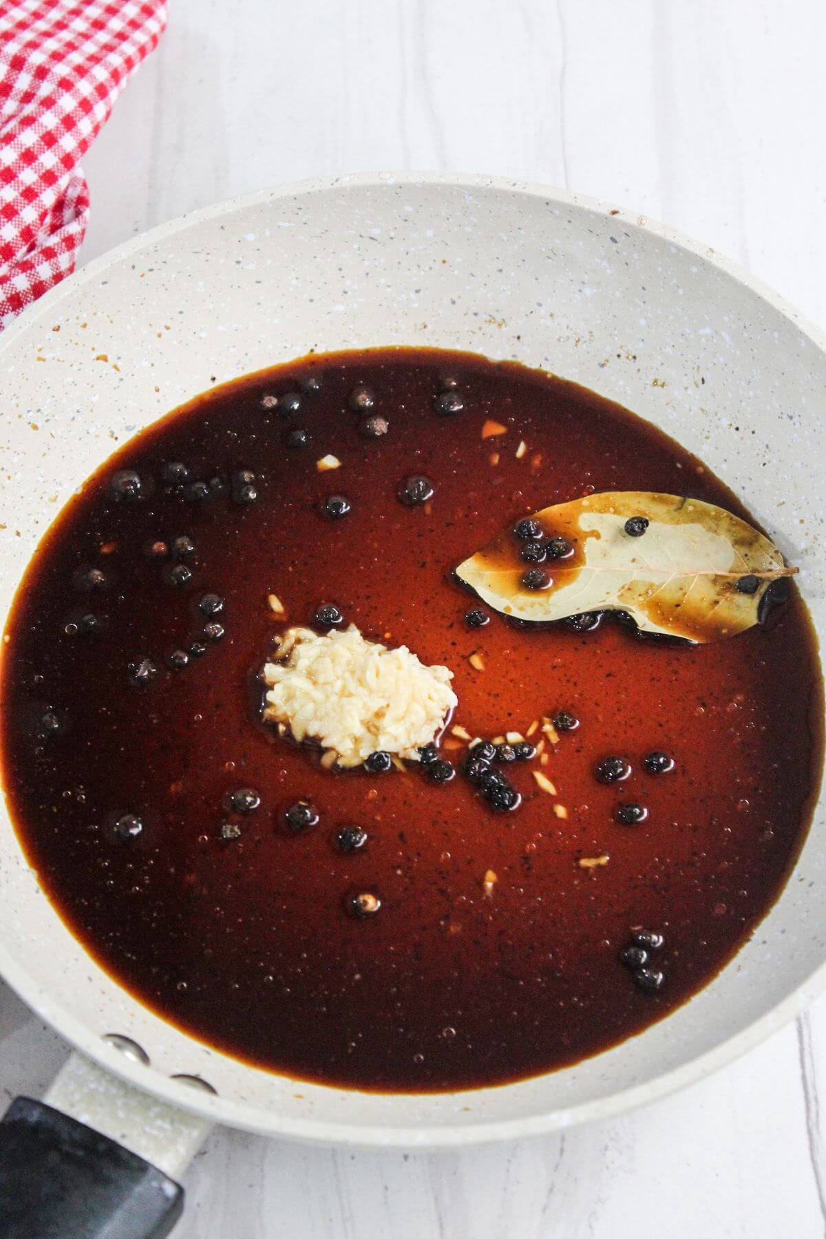 A pan containing a dark brown liquid mixture with whole spices, a bay leaf, and a dollop of minced garlic. A red and white checkered cloth is partially visible on the side.
