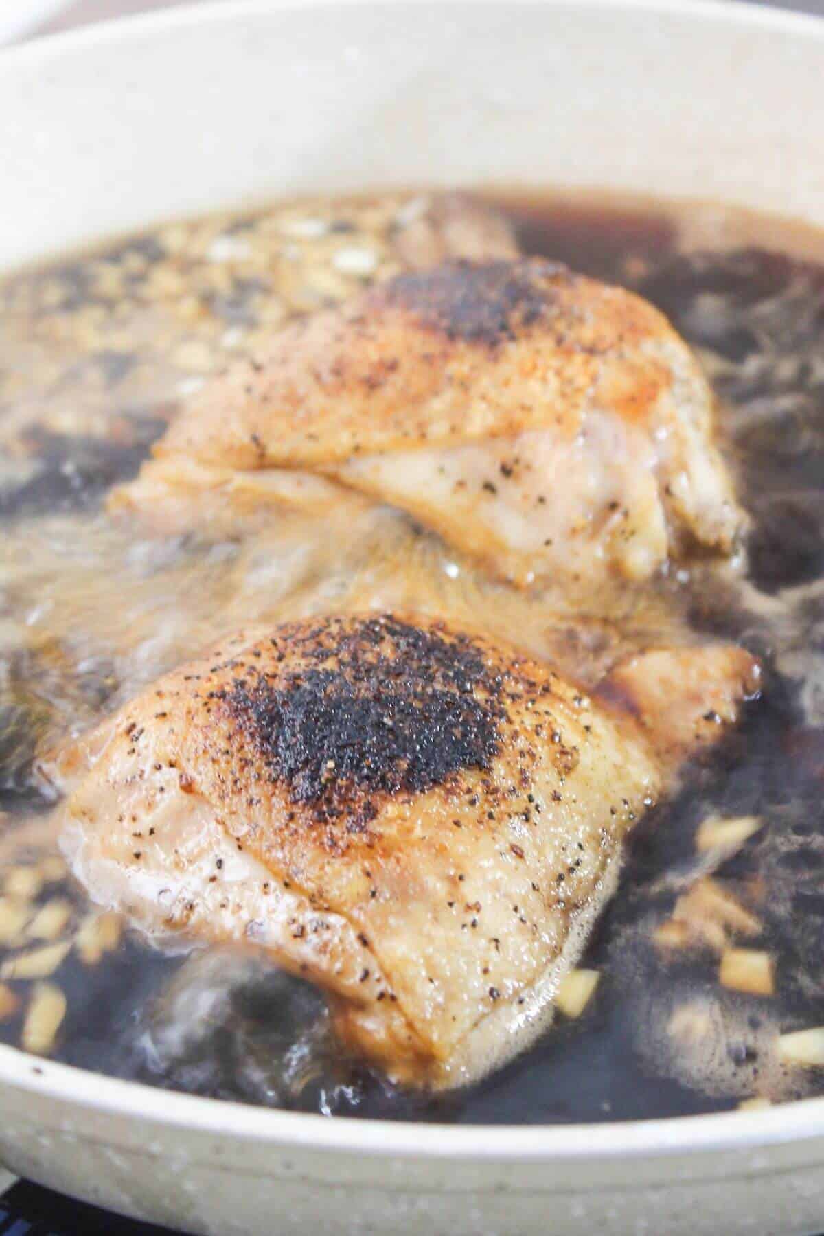 Two seasoned chicken thighs are simmering in a pan with a dark sauce and chopped garlic.