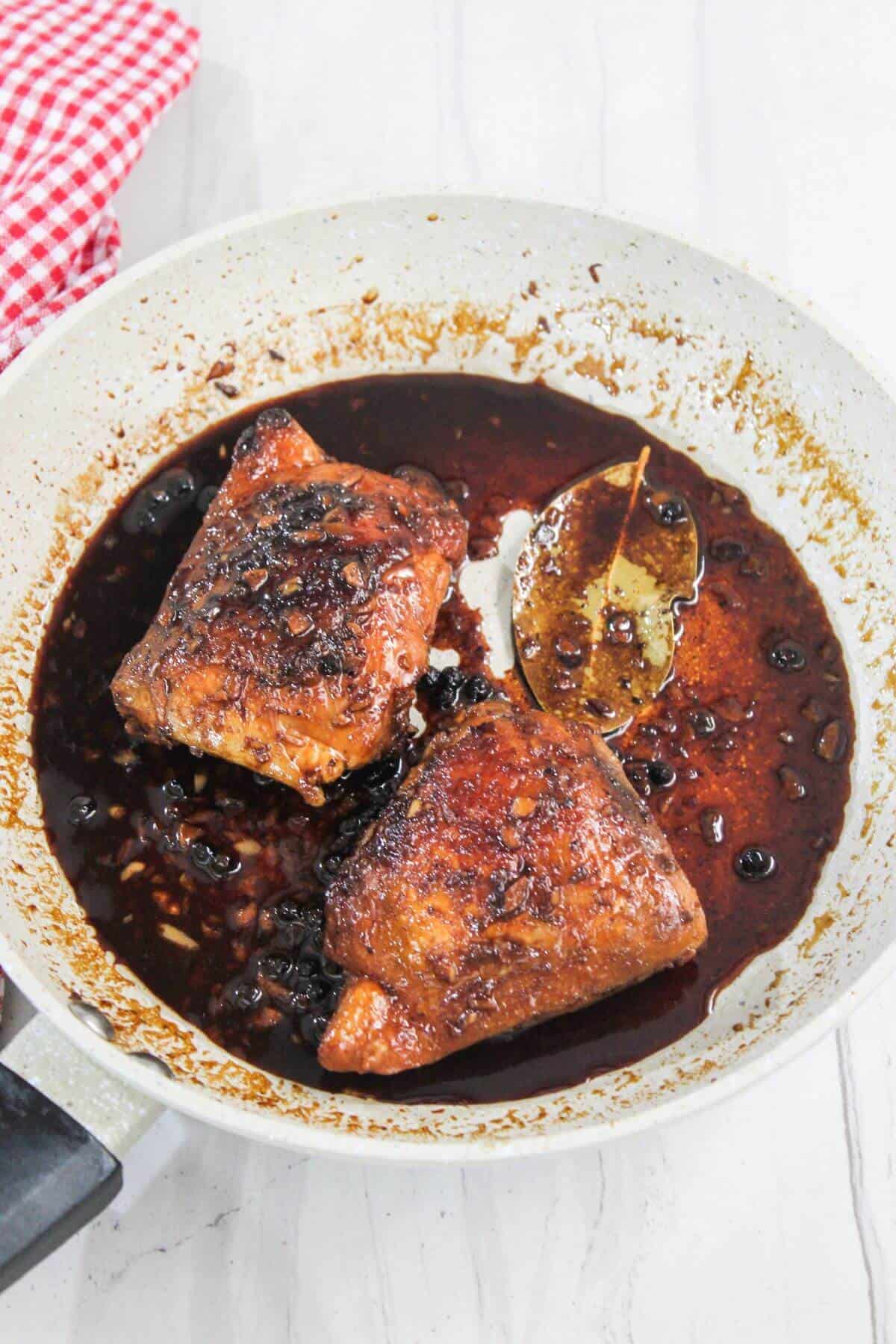 Two pieces of chicken in a pan with dark sauce, featuring a visible bay leaf. A red and white checkered cloth is partially visible.