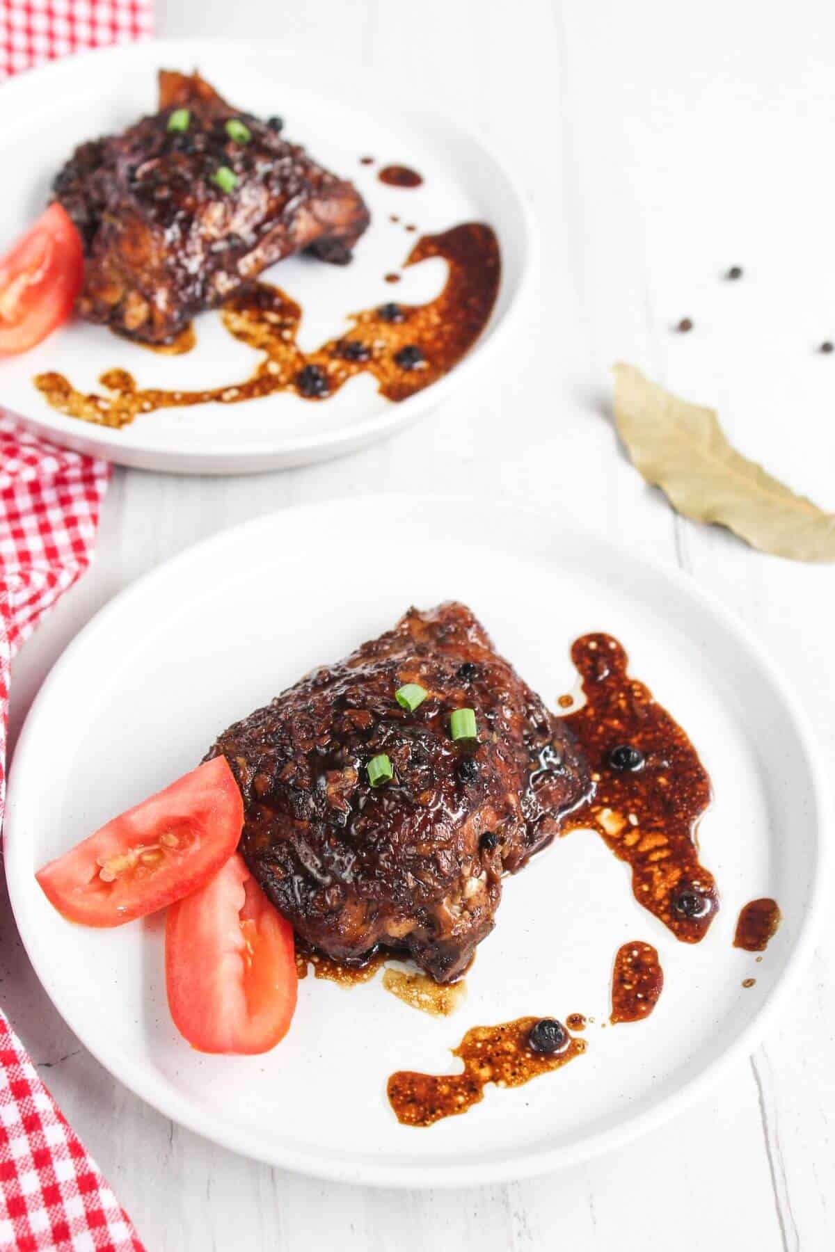 Two plates of glazed chicken with black pepper sauce, garnished with chopped green onions and tomato slices, on a white table with a red checkered cloth.