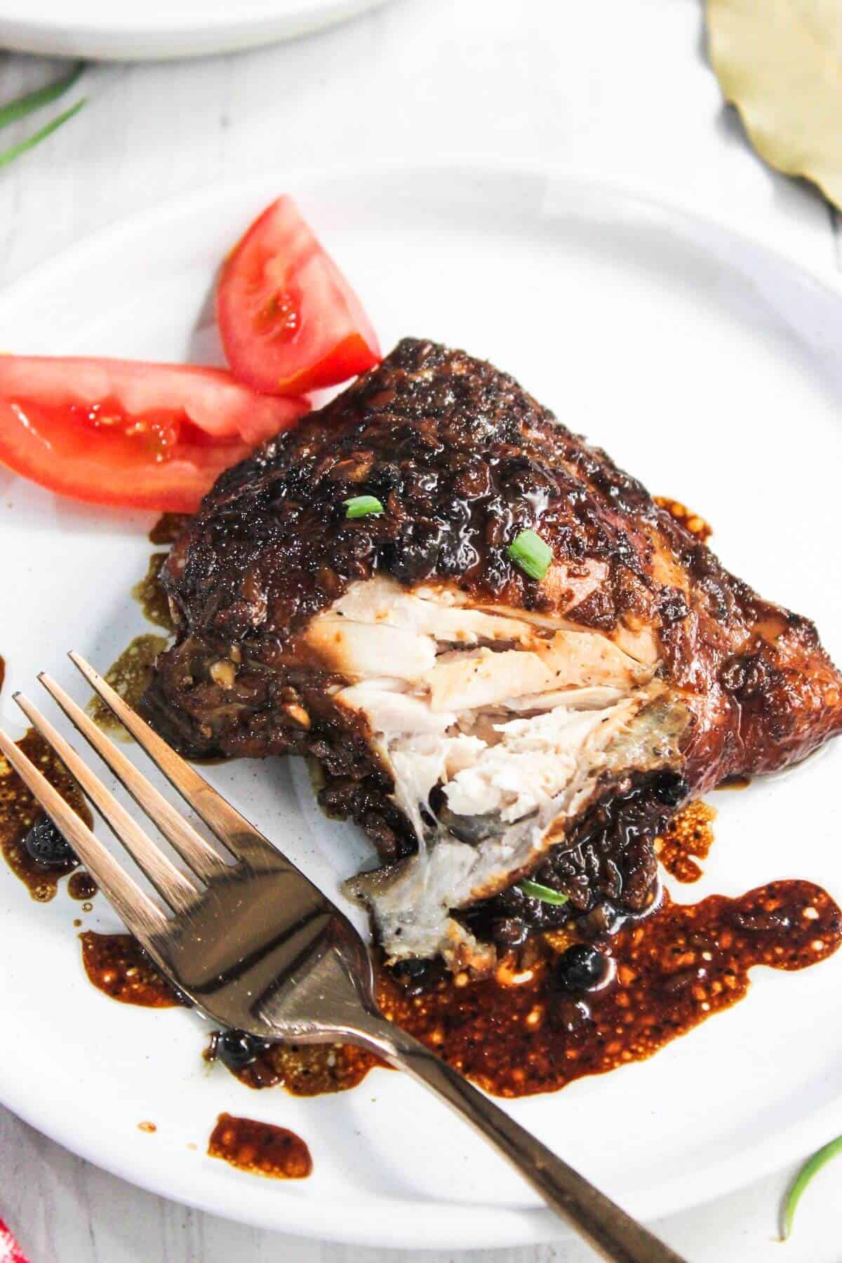 A piece of glazed grilled chicken with a fork on a white plate, garnished with tomato slices and sauce.