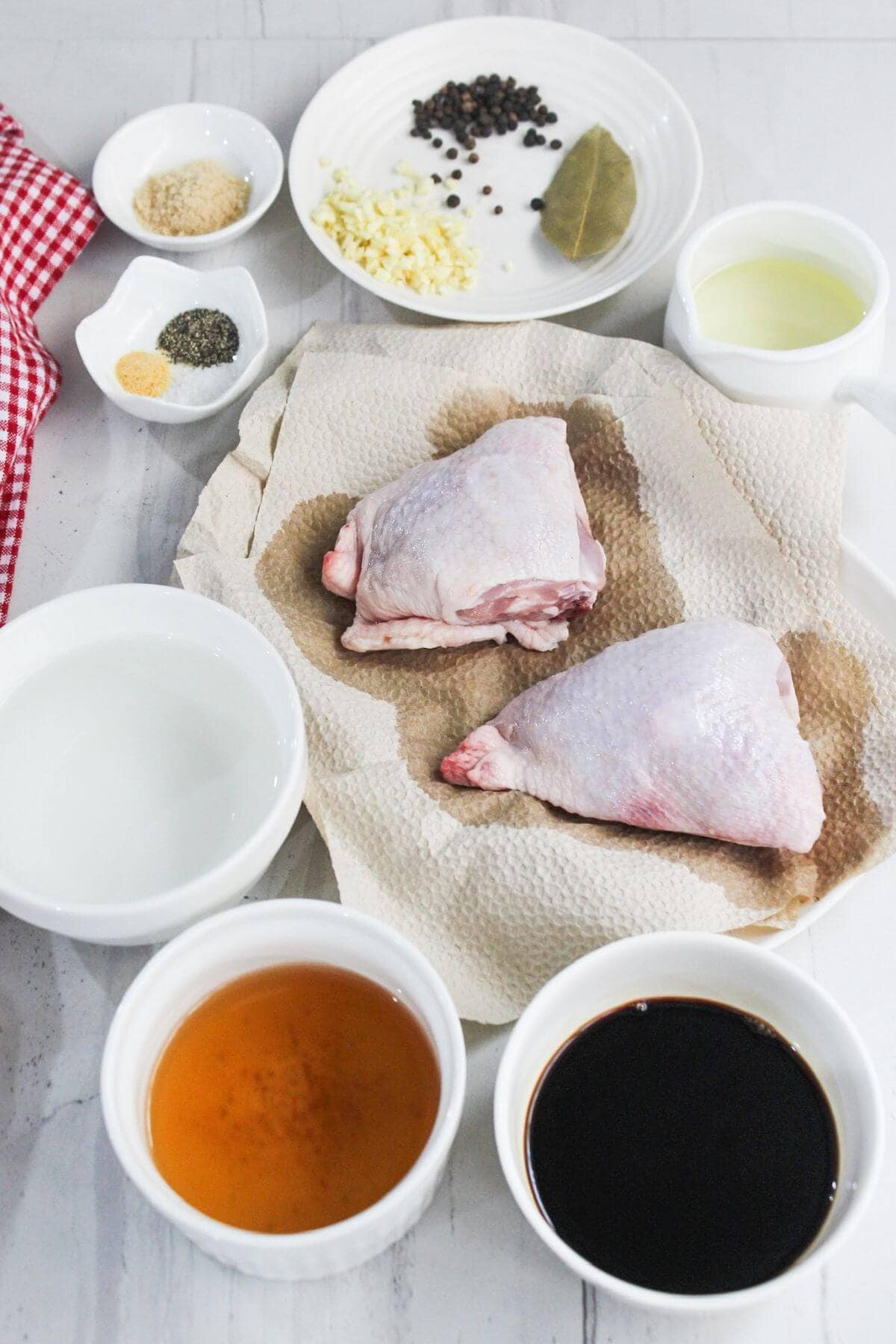Ingredients arranged for a recipe: two raw chicken thighs on a paper towel, bowls of soy sauce, vinegar, oil, garlic, peppercorns, bay leaf, and spices on a countertop.