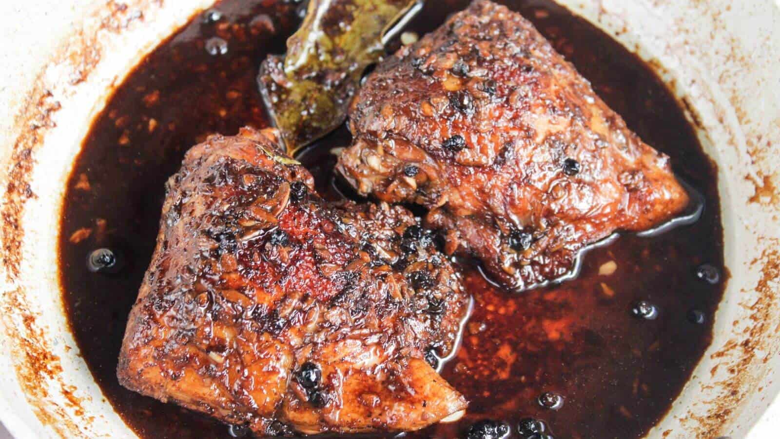 Two pieces of braised meat in a dark, rich sauce, seasoned with whole peppercorns and a bay leaf, in a pan.