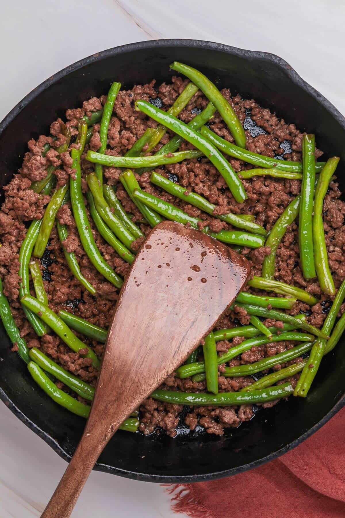 A wooden spatula in a skillet with cooked ground beef and green beans.