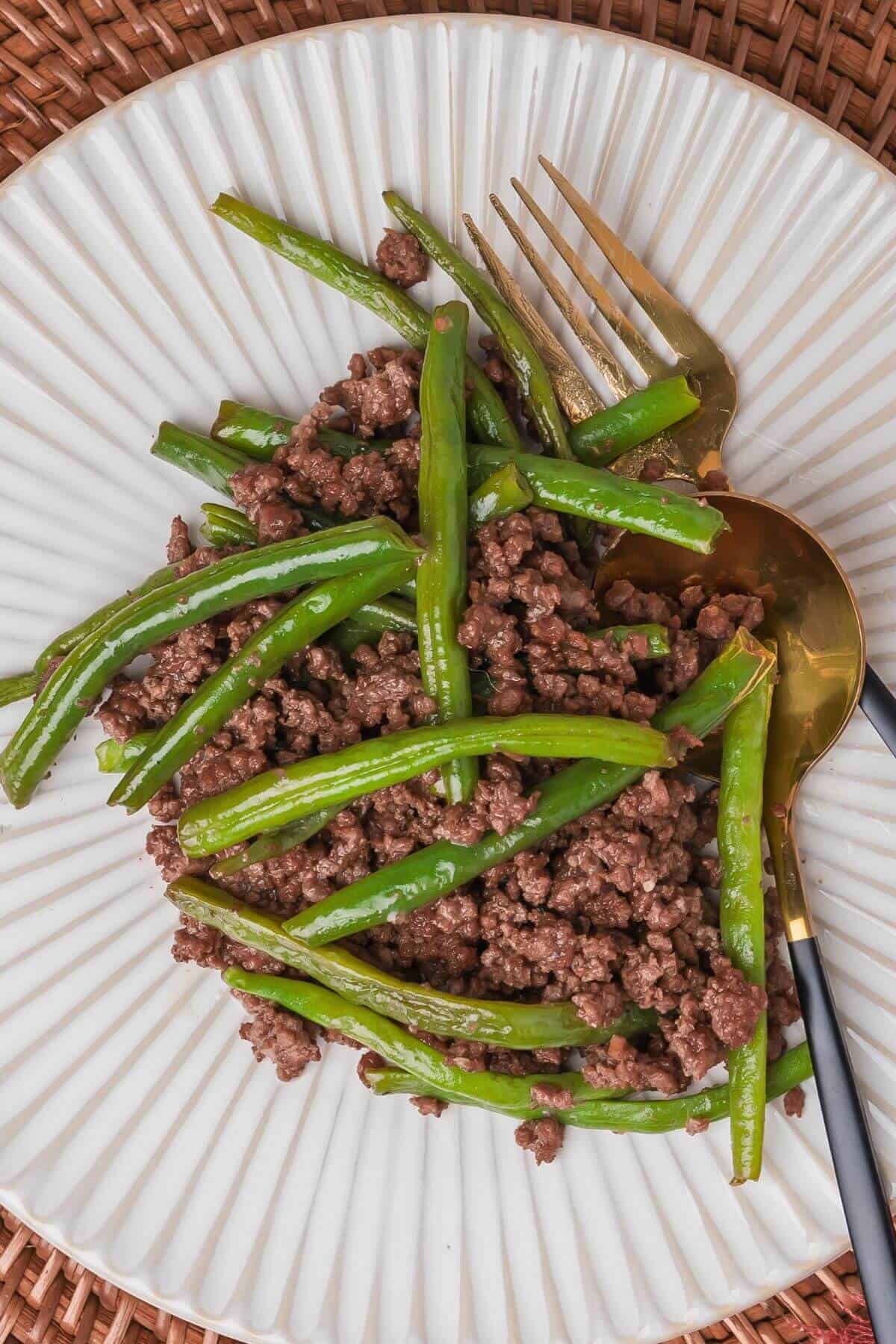 Plate of cooked ground beef mixed with green beans, alongside a fork and spoon on a textured white plate.