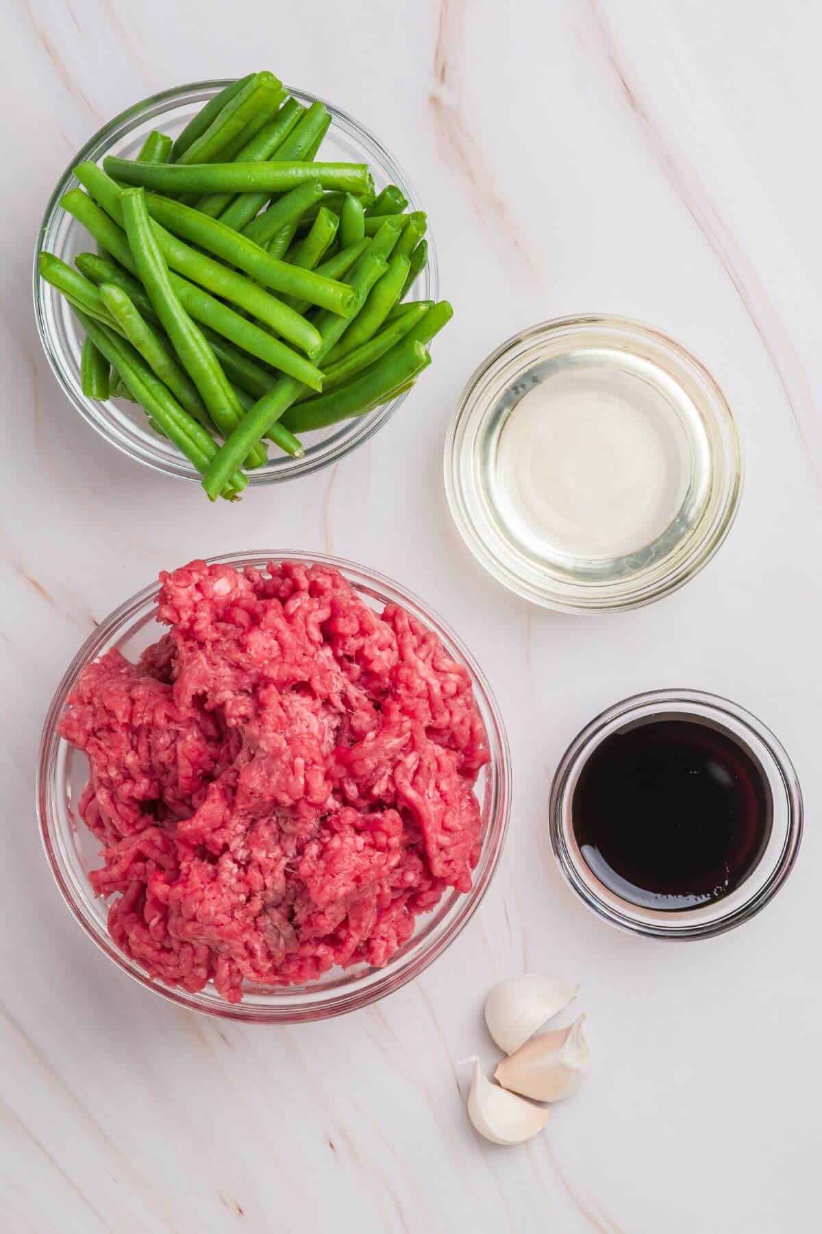 Bowls with raw ground meat, green beans, a clear liquid, and a dark sauce arranged on a marble surface. Two garlic cloves are nearby.