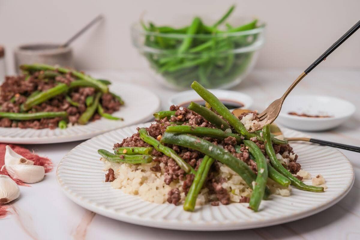Plates of stir-fried green beans and ground meat on rice with a bowl of raw beans in the background, garnished with garlic cloves and sauce.